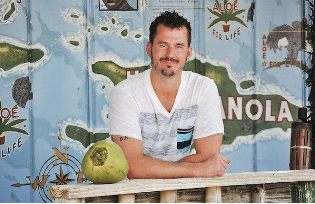 A man with short dark hair and a beard sitting at a wooden counter with a green coconut in front of him, smiling and wearing a white t-shirt with a pocket. Behind him is a colorful mural of a map of the Florida area, including the words 'Aloe for Life' and 'NOLA'.