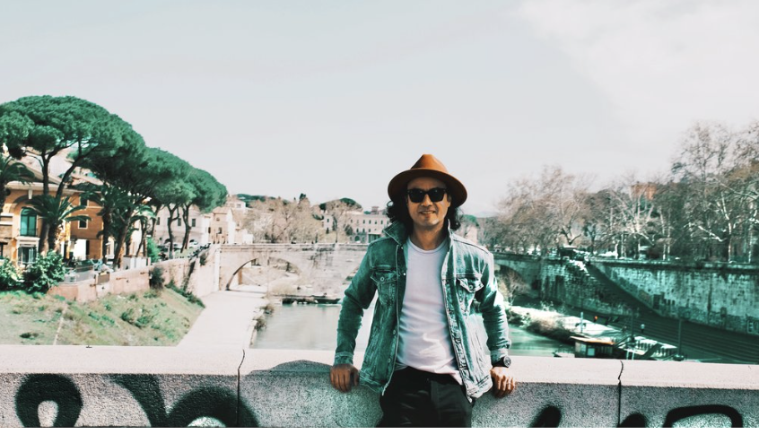 Person wearing a wide-brimmed hat, sunglasses, and denim jacket standing by a stone railing along a river with trees and buildings in the background.