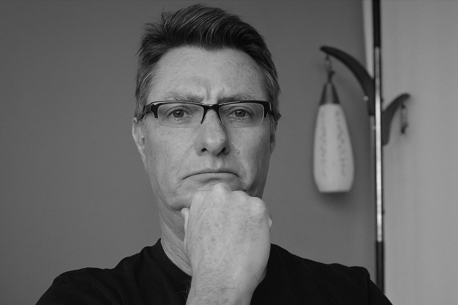 Black-and-white photo of a middle-aged man with glasses, thoughtful expression, hand on chin, standing in front of scales in a room.