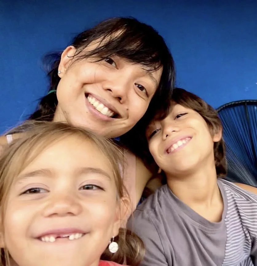 A woman with dark hair smiling with two children, one girl and one boy, all showing missing front teeth and smiling. They are indoors against a blue background.