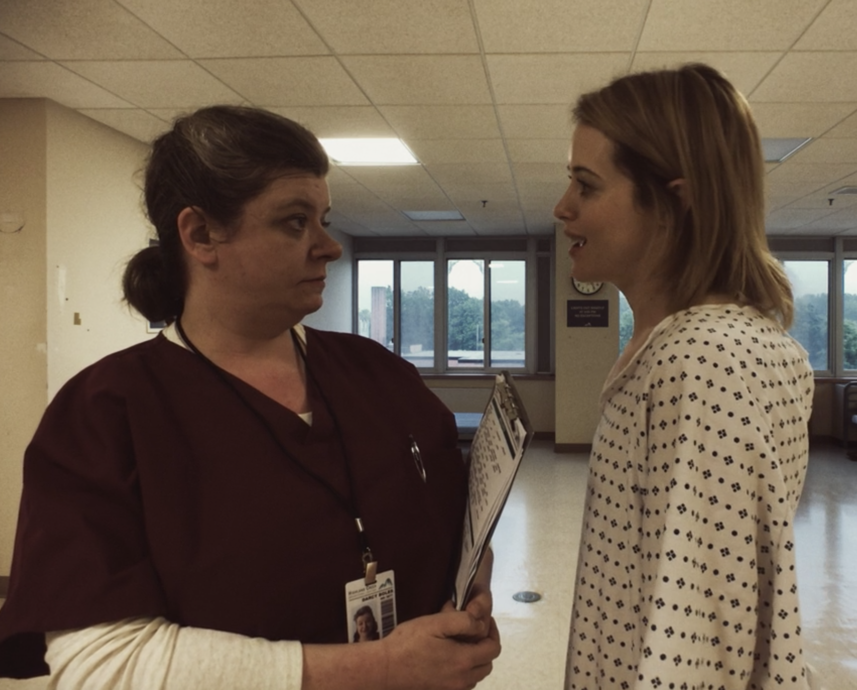 A nurse or medical worker in burgundy scrubs holding a clipboard with an ID badge, standing face to face with a woman in a white polka-dot blouse in a hospital or medical facility hallway with large windows in the background.