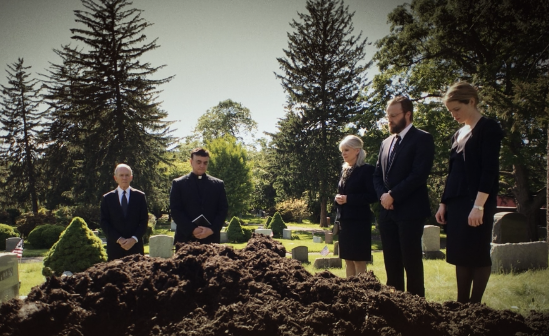 People standing in a cemetery during a burial or memorial service, with a large mound of dirt in the foreground and trees behind them.