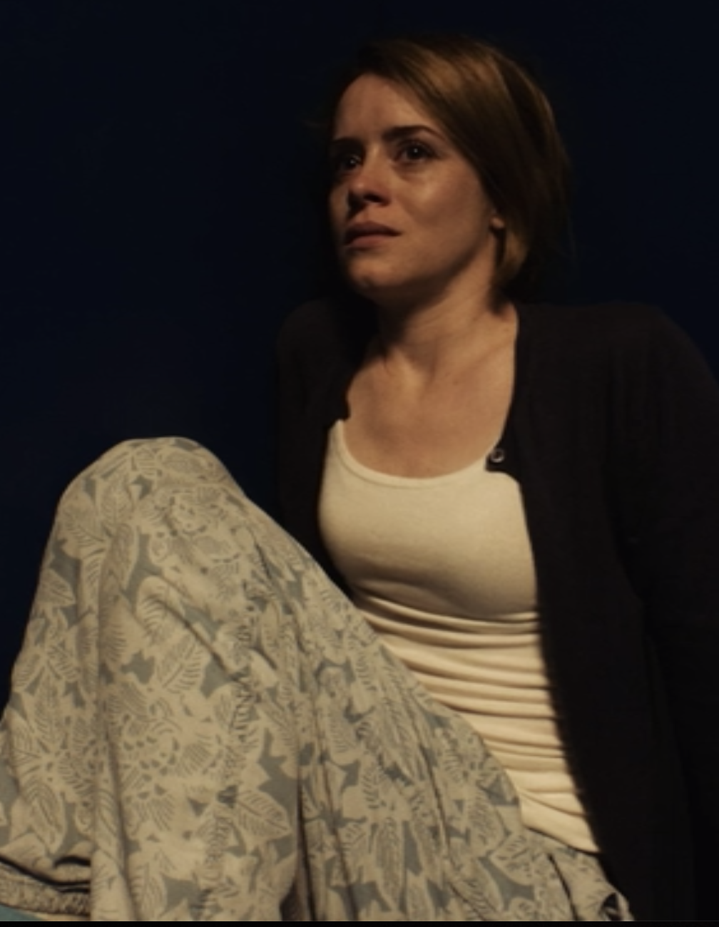 A woman with short brown hair, wearing a black jacket and white shirt, sitting against a dark background.