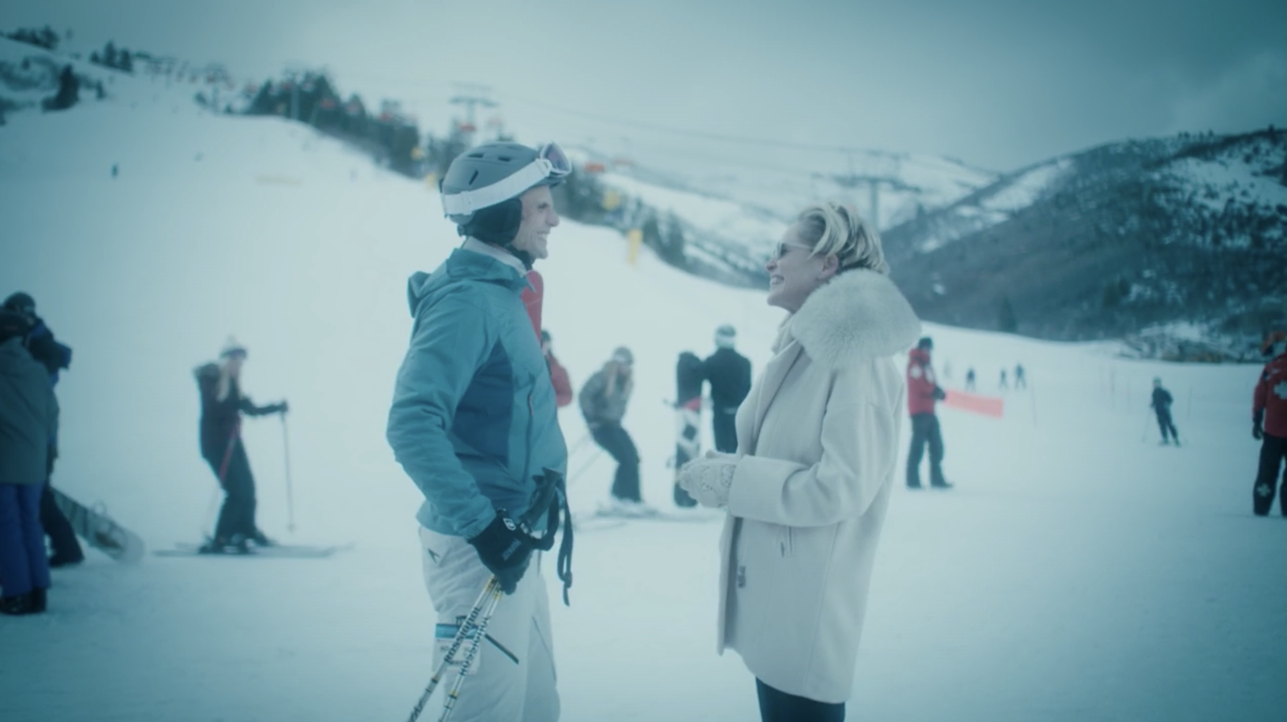 A man and woman smiling and talking on a snowy ski slope, with other skiers in the background and ski lifts on a mountain.