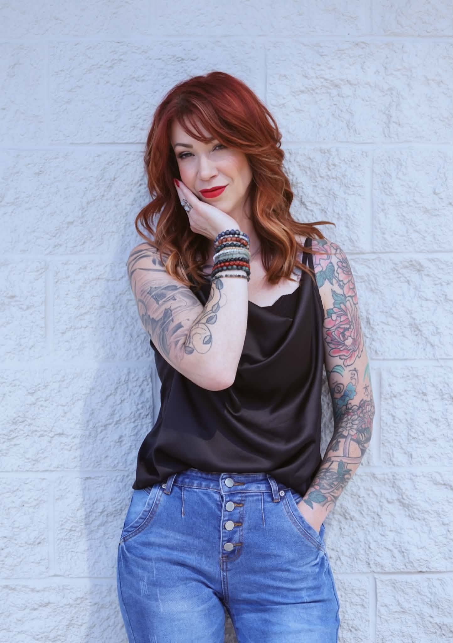 Woman with tattoos, red hair, and colorful bracelets standing against a white brick wall, winking and resting her face on her hand.