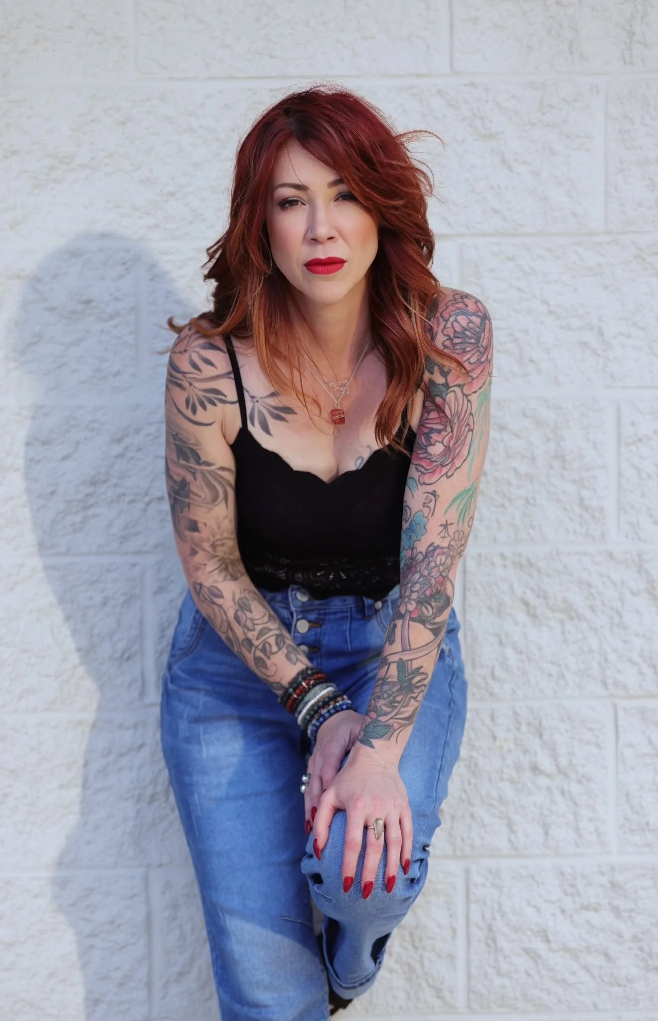 A woman with red hair, tattoos on her arms, wearing a black top and blue jeans, standing against a white brick wall, winking, and holding her hands in front of her.