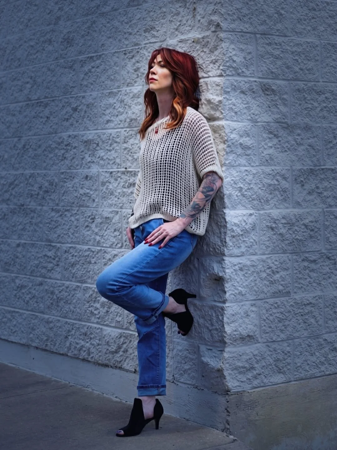 Woman with red hair and tattoos leaning against a textured gray brick wall, wearing a white crochet top, blue jeans, and black high heels.
