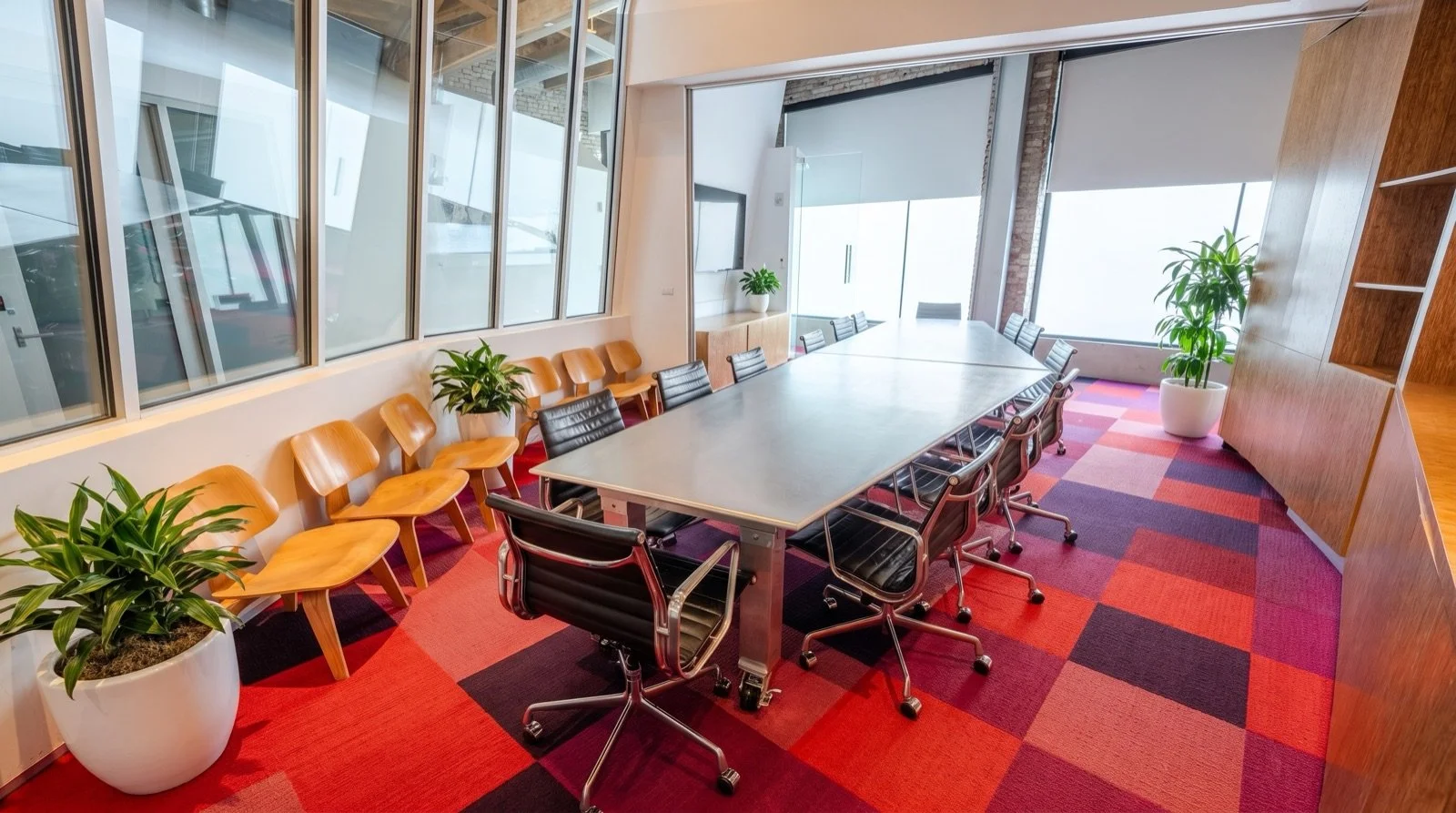 Modern conference room with a large central table, office chairs, potted plants, and large windows letting in natural light.