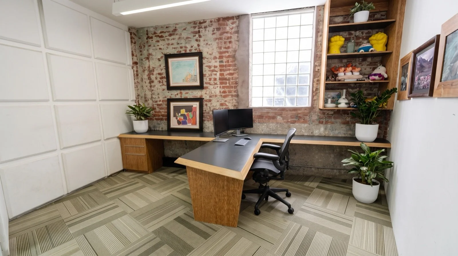 Office space with a black desk, computer monitor, office chair, two potted plants, artwork on a brick wall, wooden shelves with decorative items, and a large window