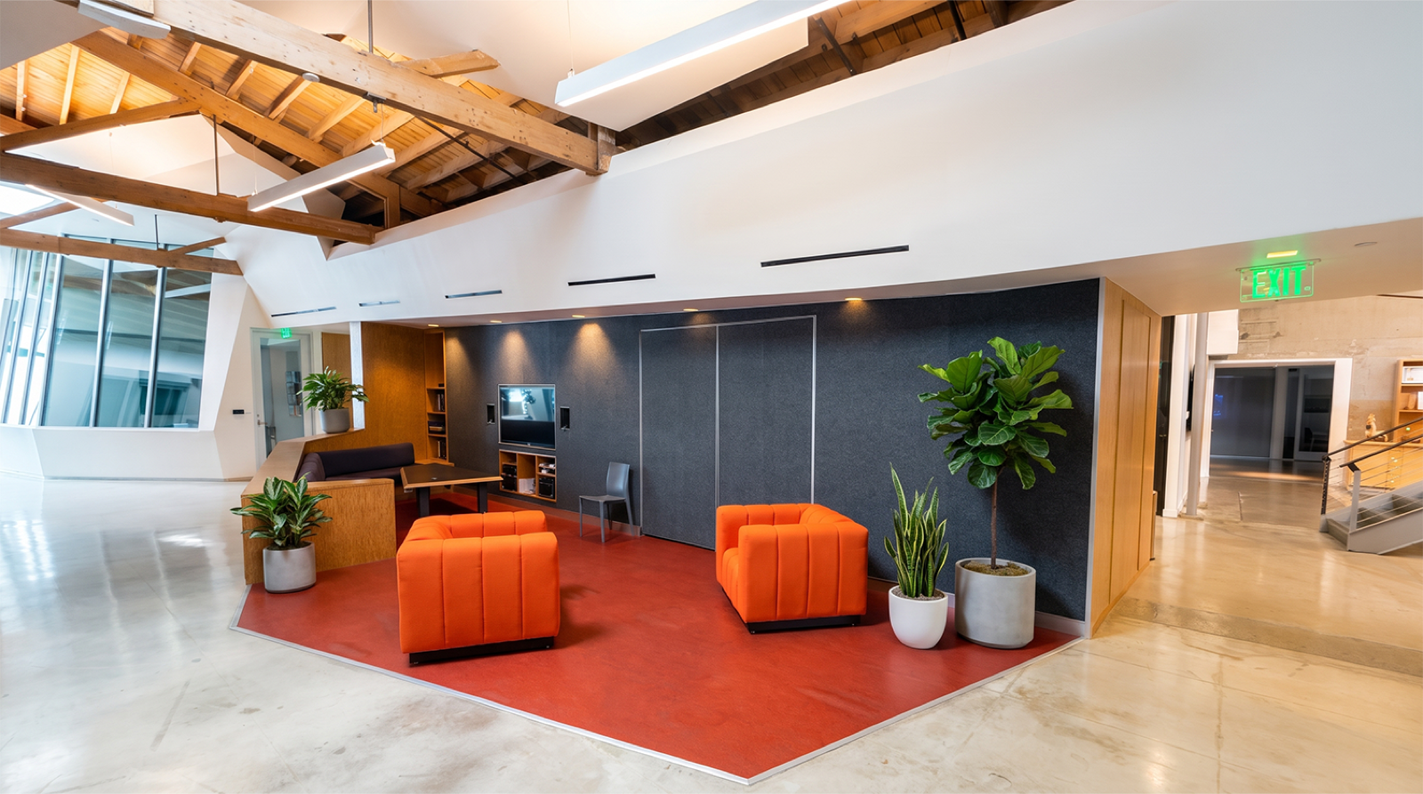 Modern office lounge area with orange chairs, a small black sofa, and a wall-mounted TV, surrounded by potted plants and illuminated by natural light and ceiling lights.