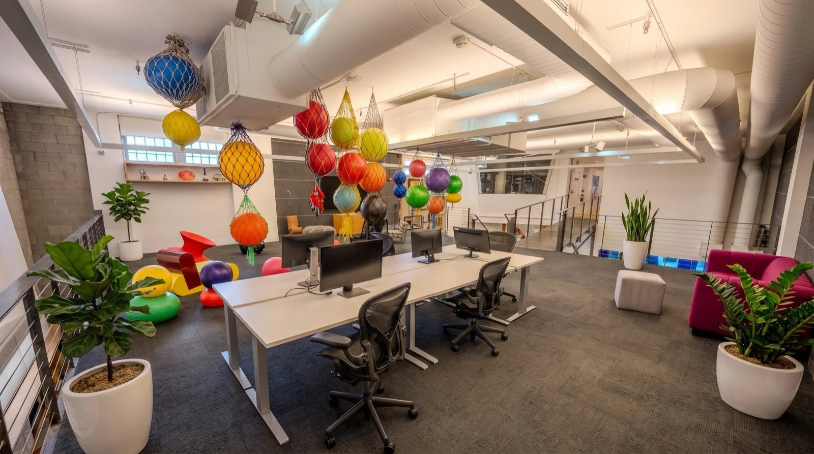 Modern office space with colorful hanging paper lanterns, potted plants, a pink sofa, and white desks with computer monitors.