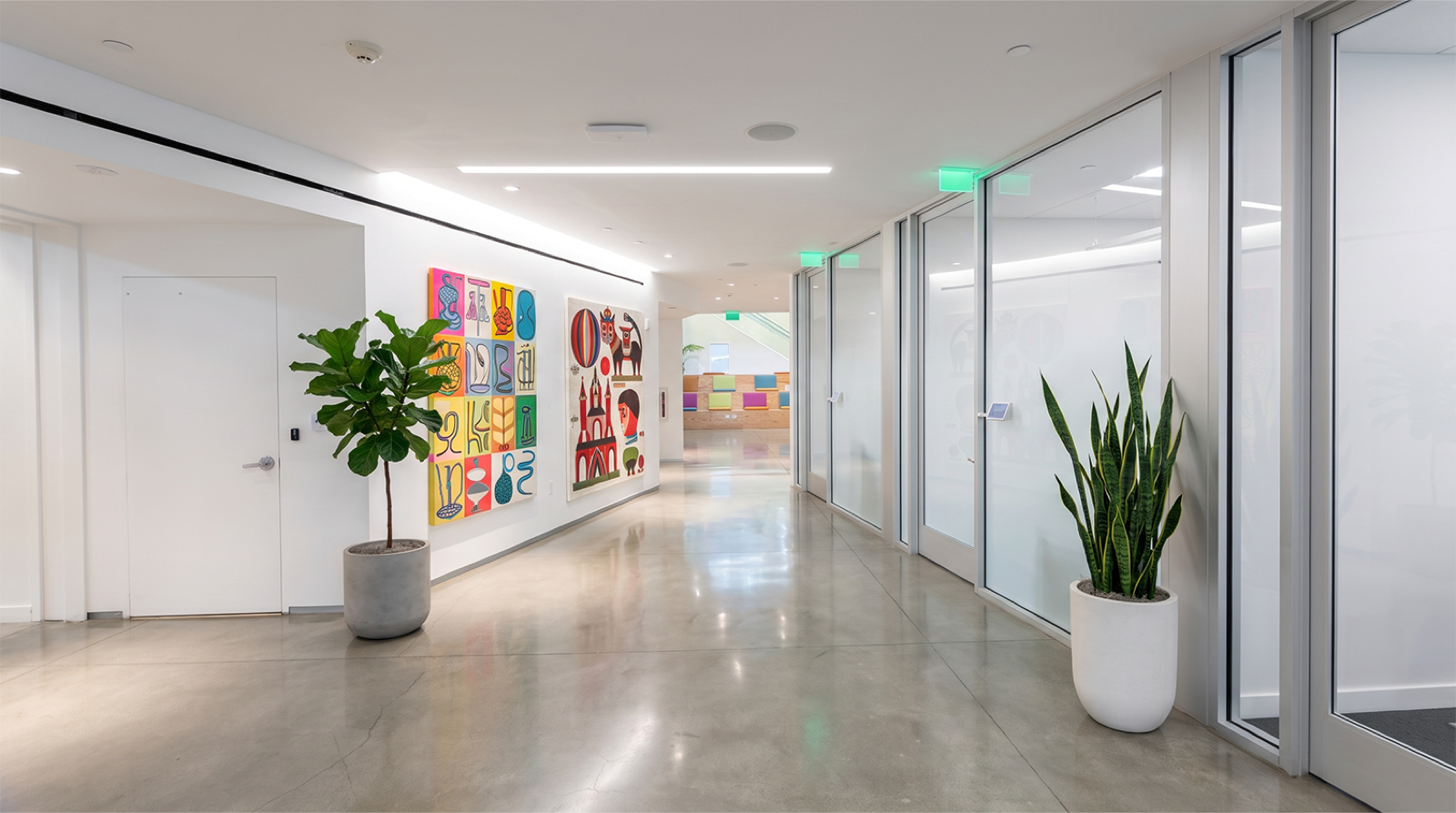Modern office hallway with abstract colorful artwork on white walls, large potted plants, glass doors, and a polished concrete floor.