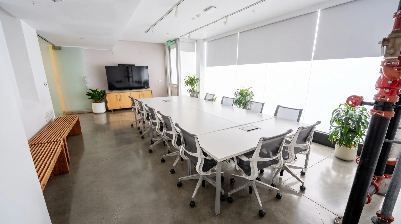 Modern conference room with a large white table, 12 office chairs, a flatscreen TV on a wooden cabinet, indoor plants, large windows with blinds, concrete floor, and minimal decor.