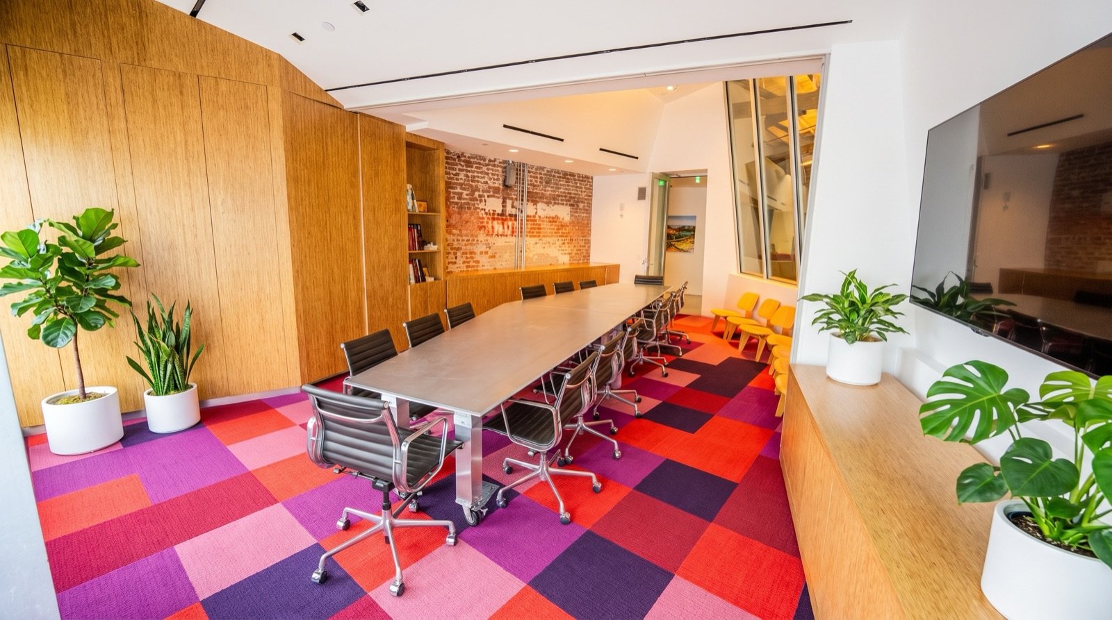 Modern conference room with a long table, black office chairs, yellow chairs, and potted plants, featuring wooden panels, brick wall, and a large TV.