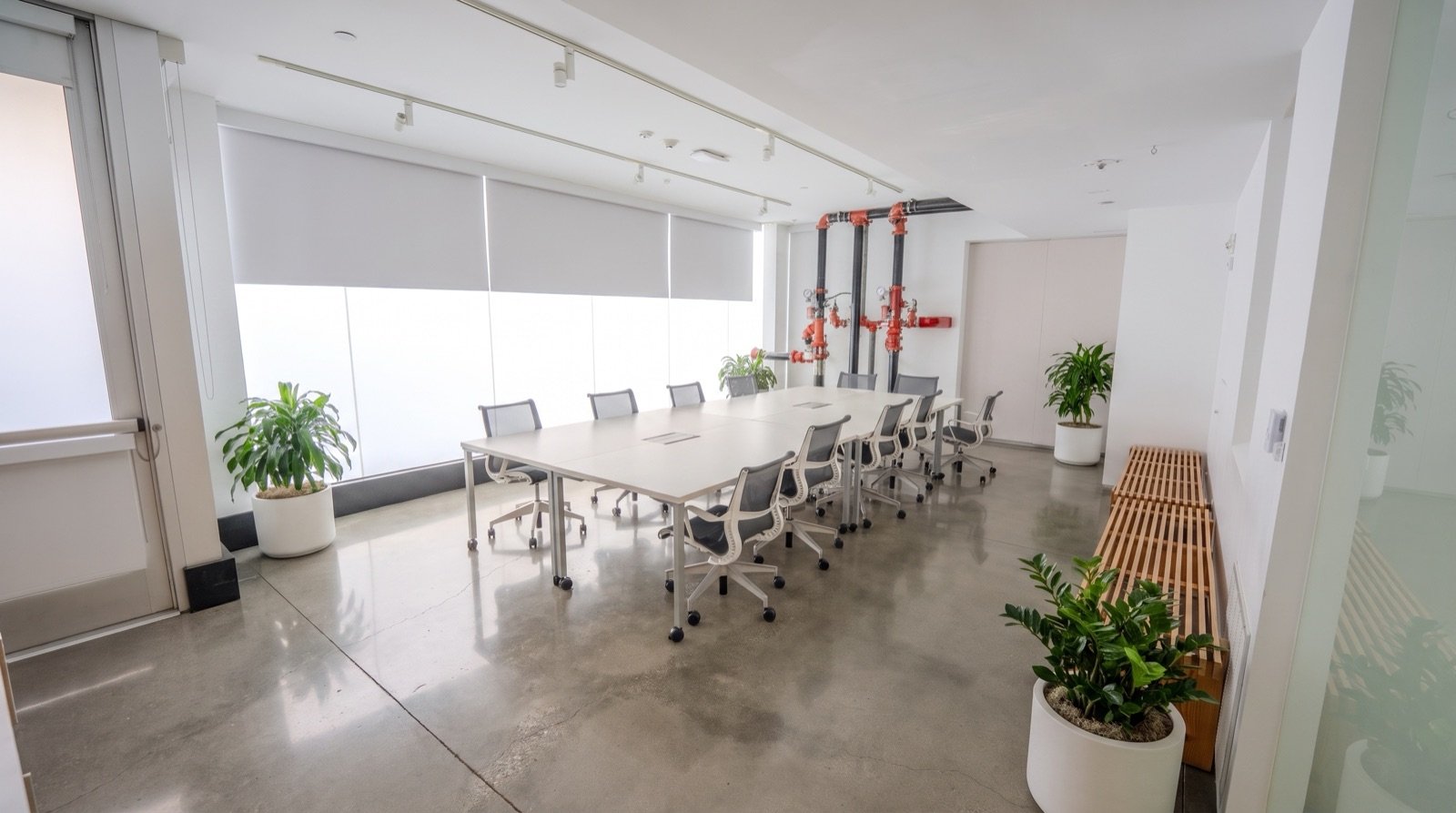 Empty modern conference room with large white table, ergonomic chairs, potted plants, and industrial piping on wall.