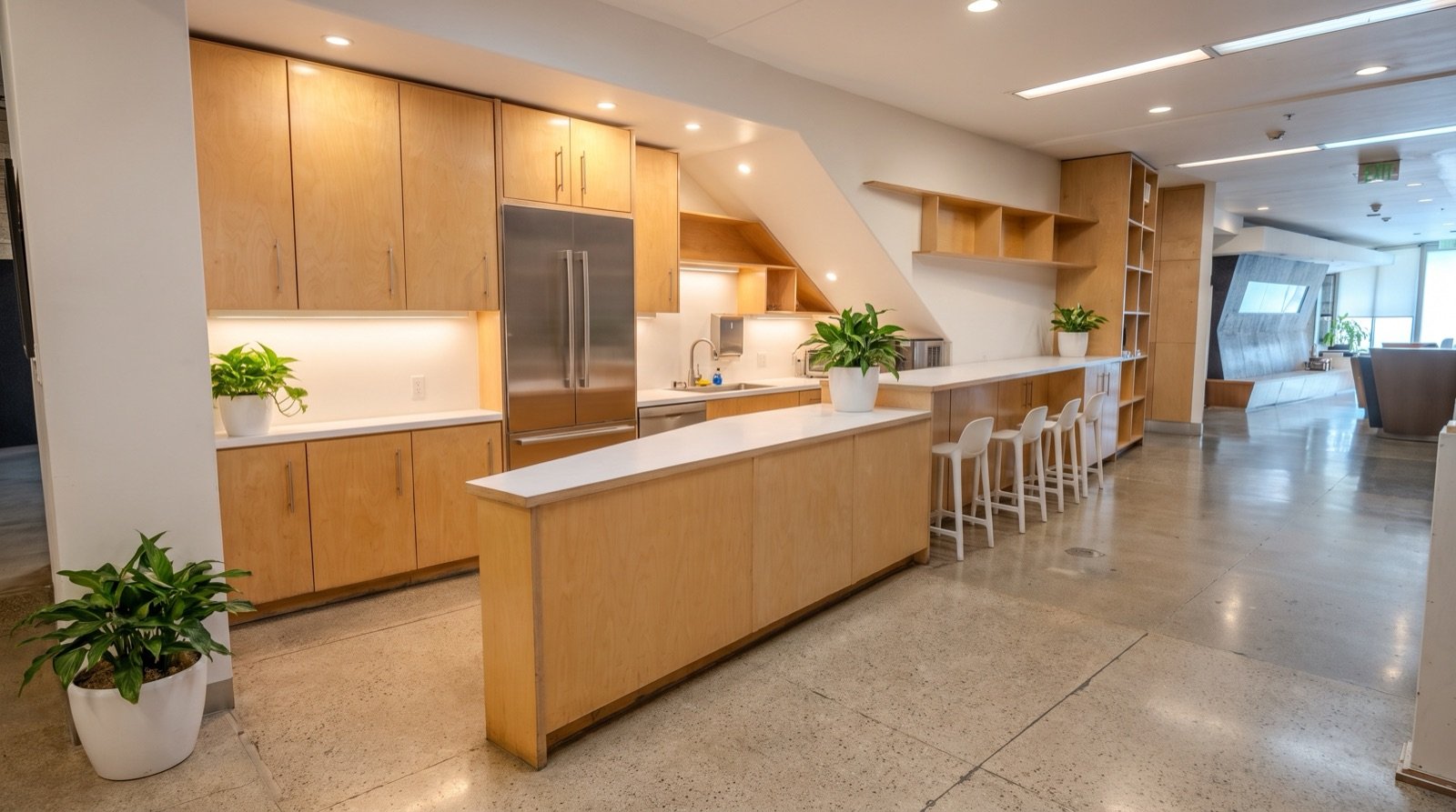Modern office kitchen with wooden cabinets, white countertops, potted plants, and bar stools, located in a bright and open office space.