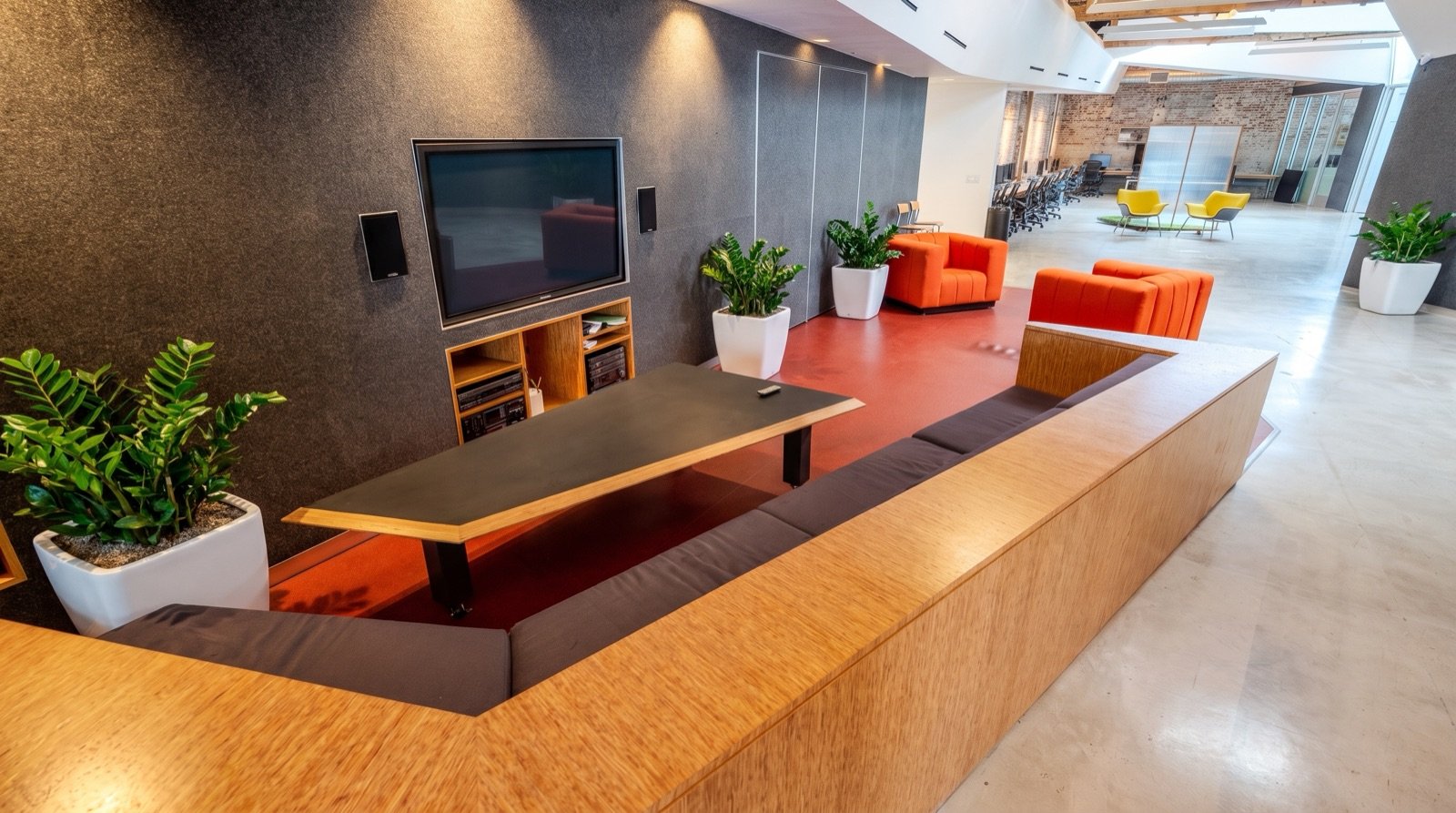 Modern office lounge area with orange and black seating, large potted plants, a flat-screen TV on a dark wall, and a conference room in the background with yellow chairs and a long table.
