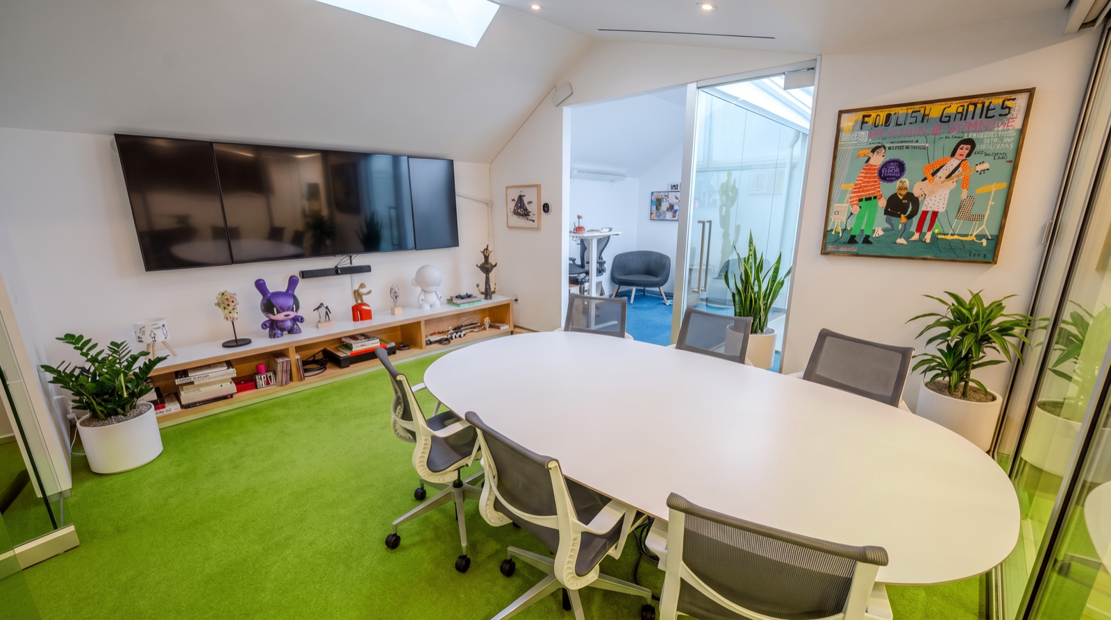 Modern office conference room with a white oval table, gray office chairs, and a bright green carpet. The wall features a large flat-screen TV, artwork, and plants. There's a glass door leading to a smaller room with more chairs and artwork.
