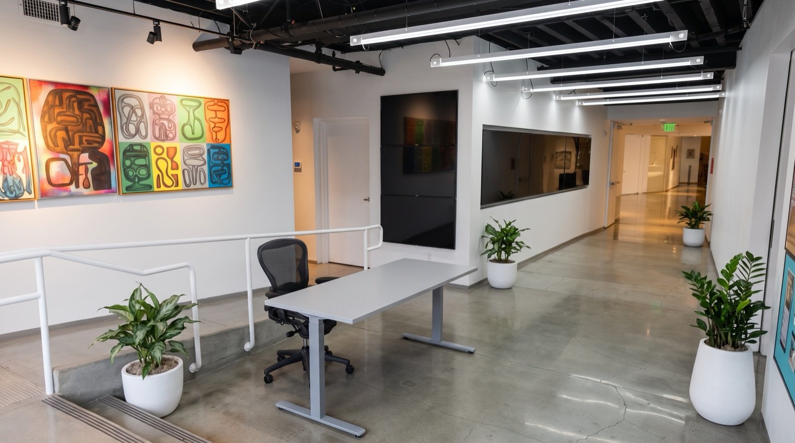 Modern office reception area with a desk, black office chair, and potted plants. Artworks on the wall and a hallway with a wall-mounted television.