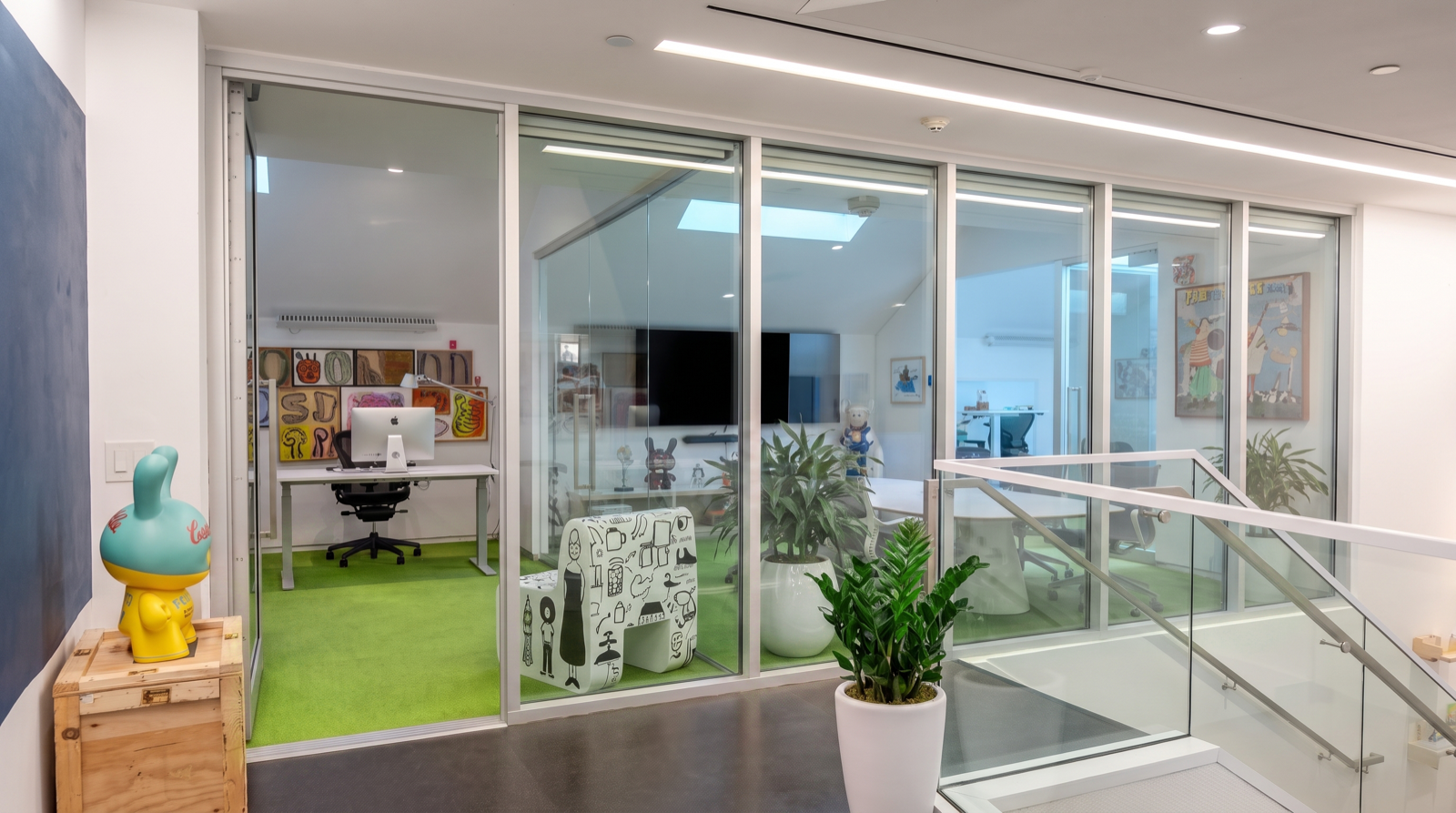 Modern office with glass walls, green carpet, art on walls, plants, and a stairway leading down.