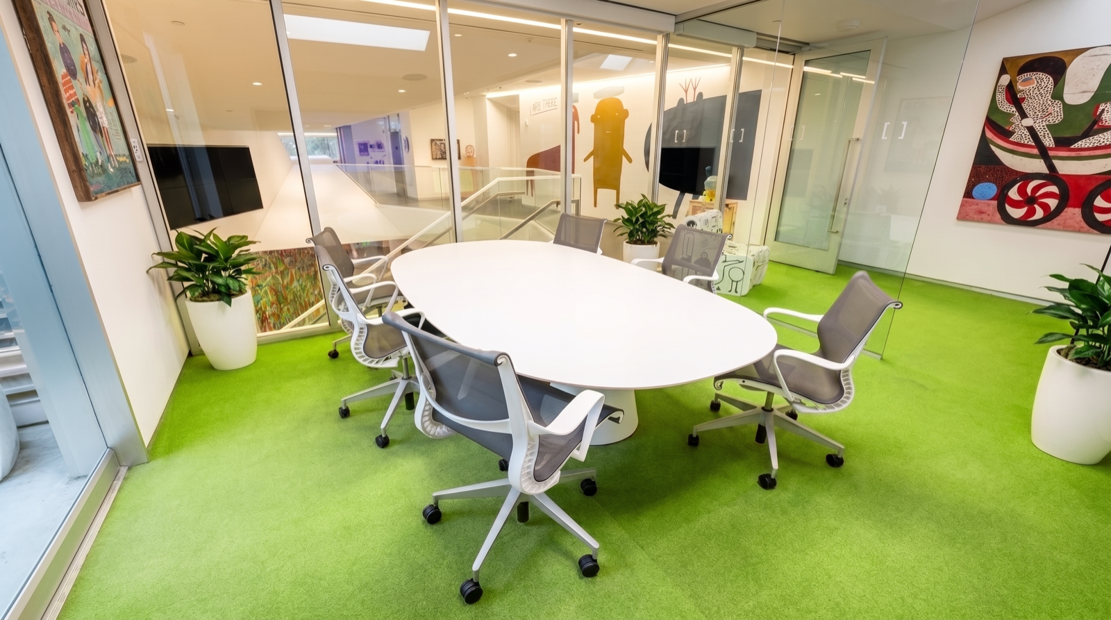 Modern conference room with green carpet, white oval table, six grey office chairs, large potted plants, glass walls with colorful artwork, and view of building interior.
