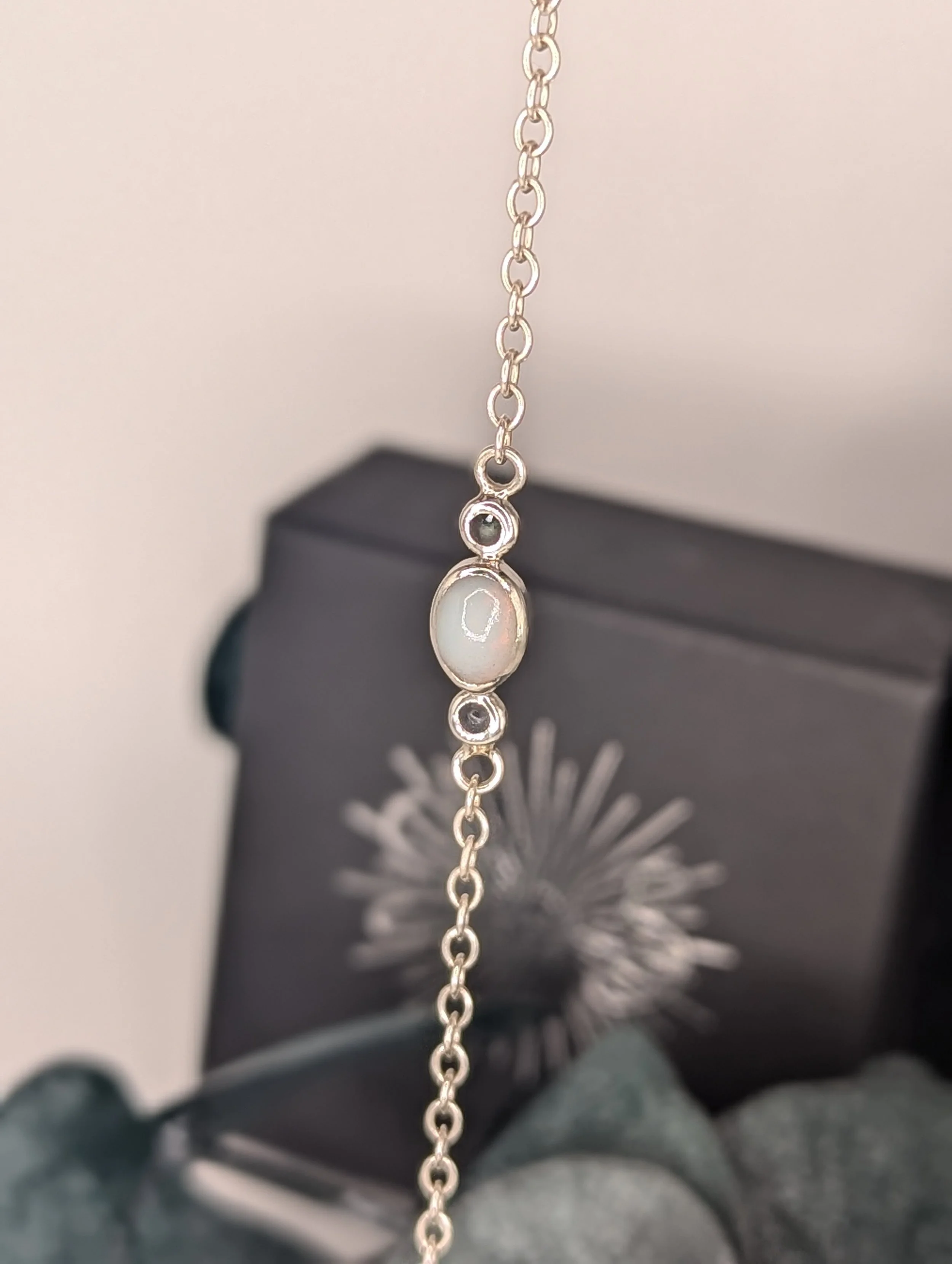 A silver necklace with a pendant featuring an oval white stone and small surrounding gemstones, held in front of a black box with a starburst design.