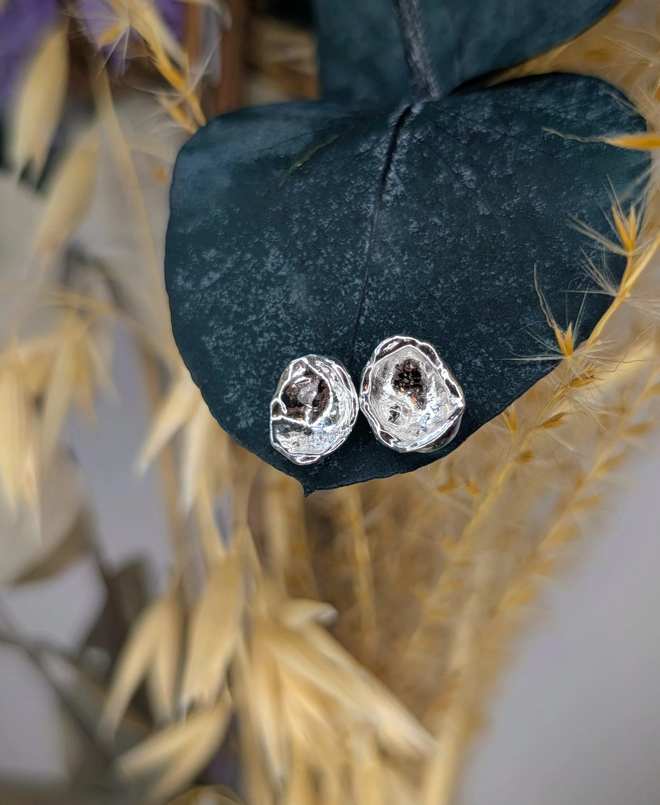 Silver earrings resting on a dark green leaf with yellowing, dried grass in the background.