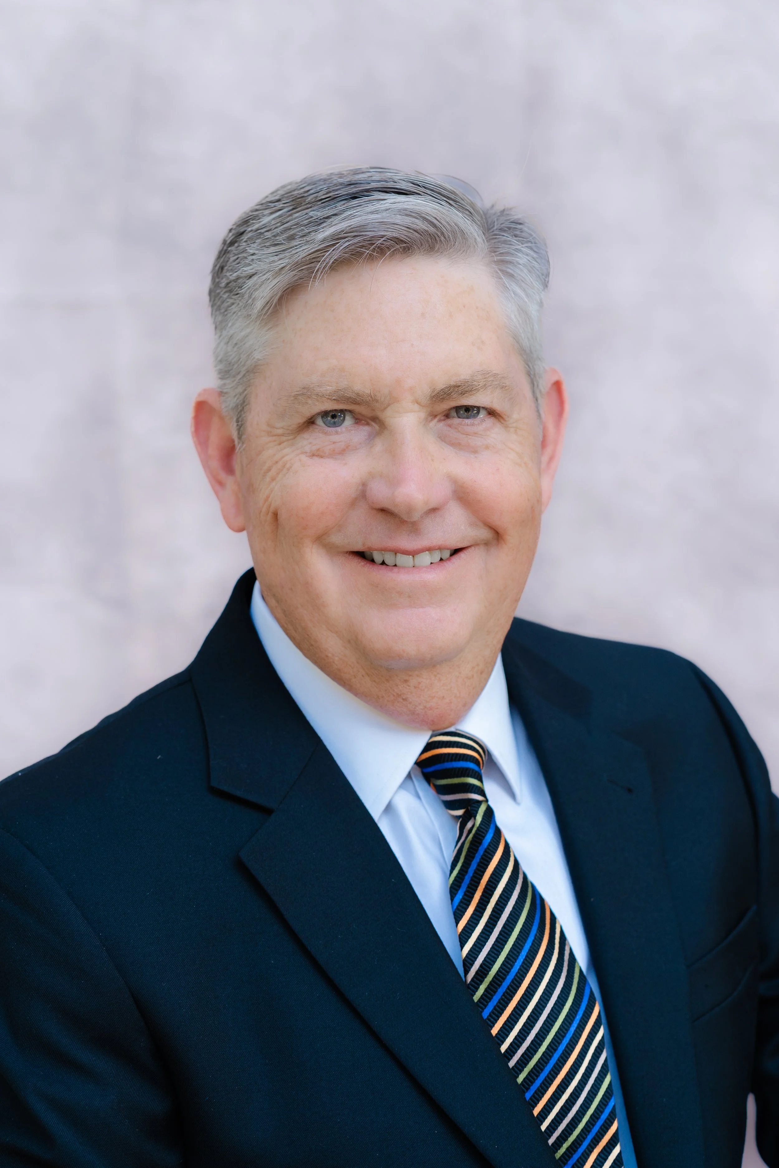 Professional headshot of a smiling man with gray hair in a navy suit and striped tie