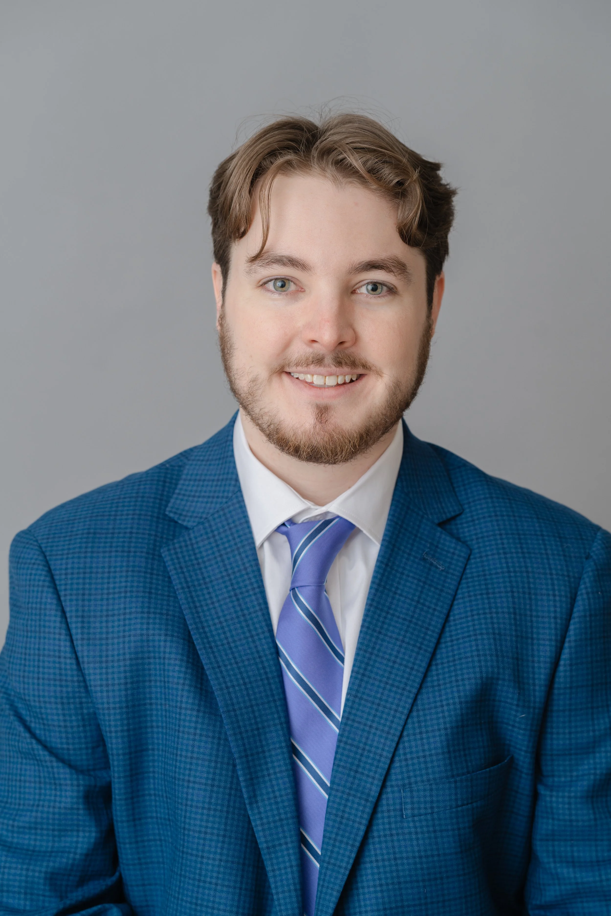 Professional headshot of a young man in a blue suit and lavender tie