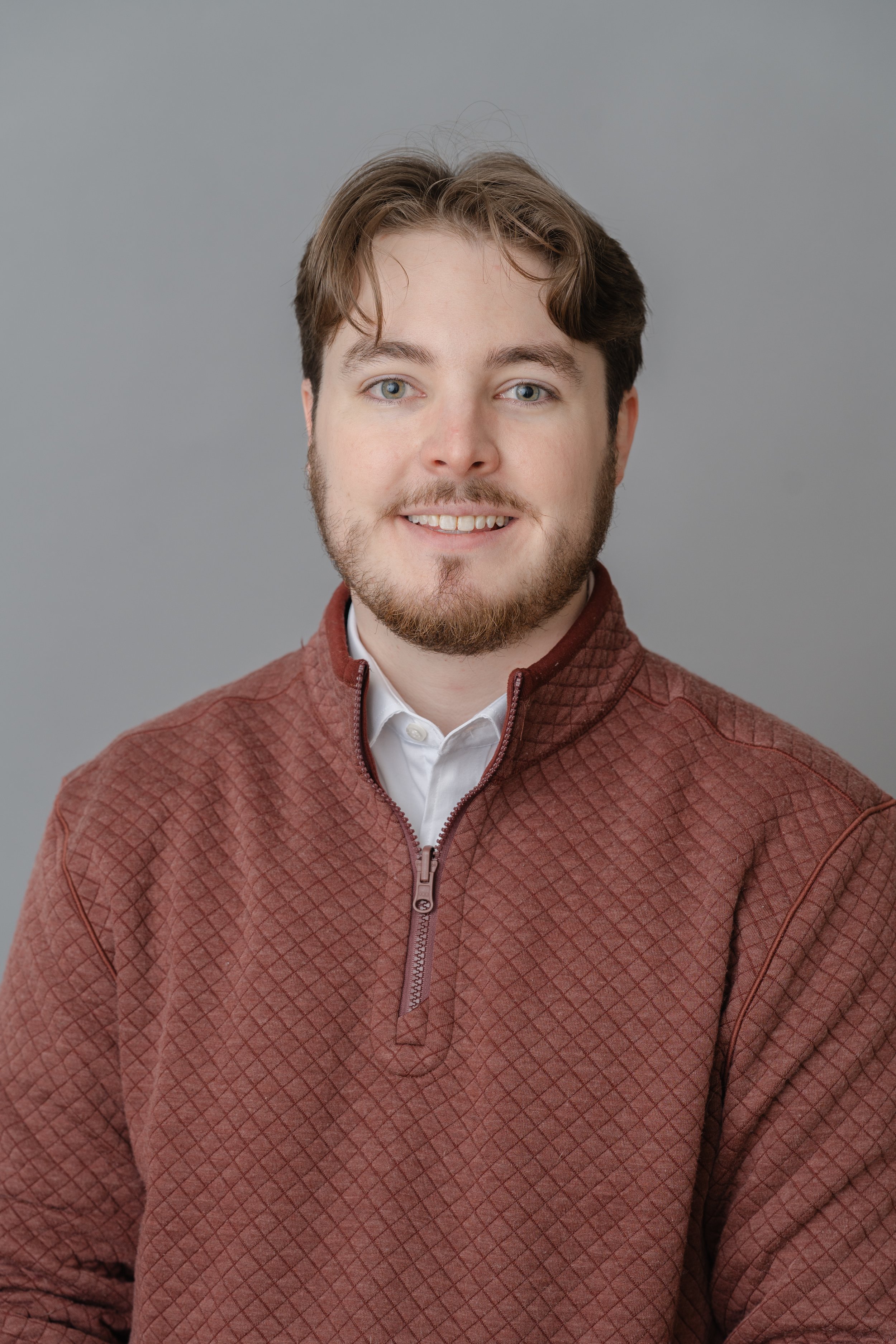 Professional headshot of a young man with a beard in a rust-colored sweater over a collared shirt