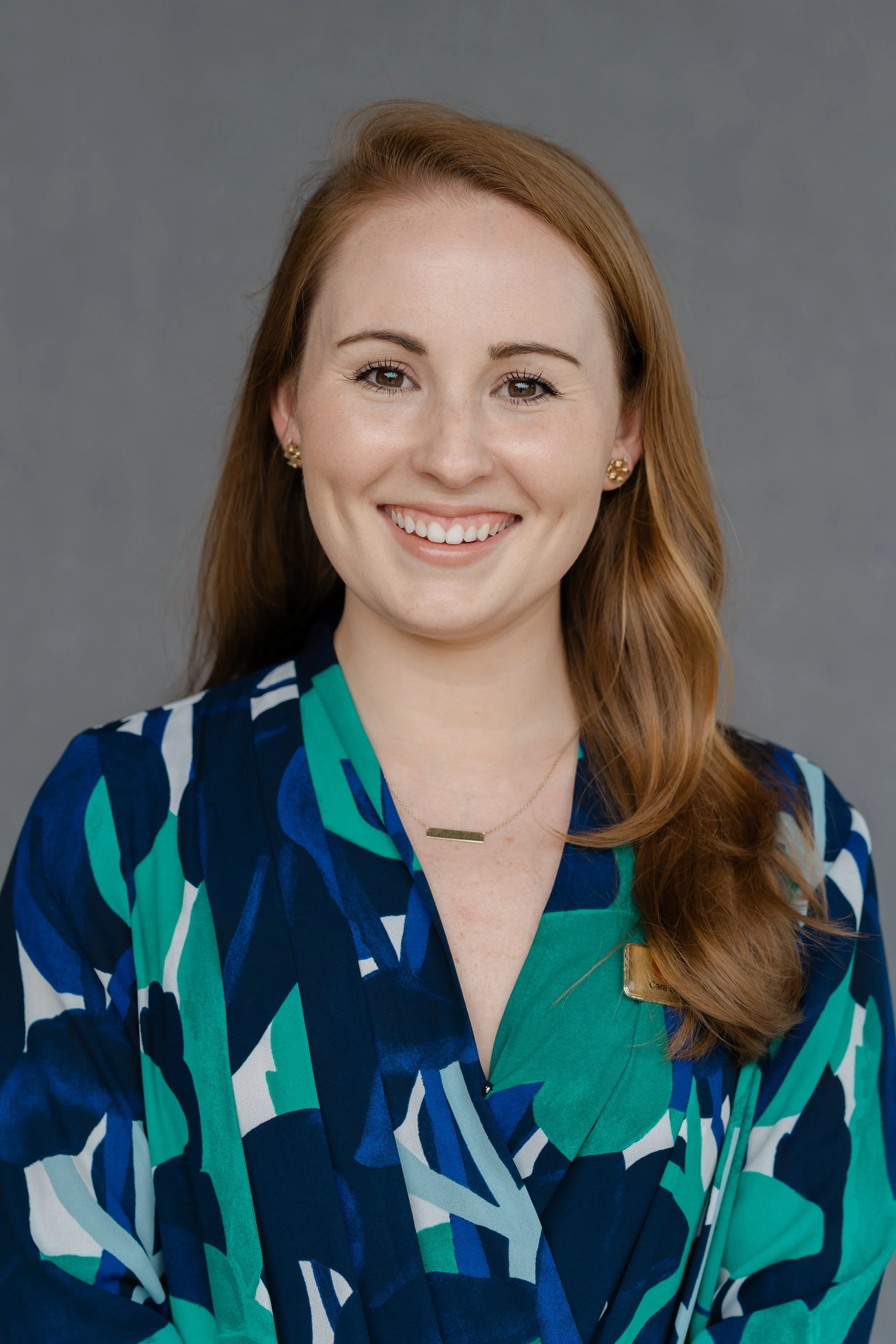 Professional headshot of a smiling woman with long auburn hair in a green and navy patterned blouse