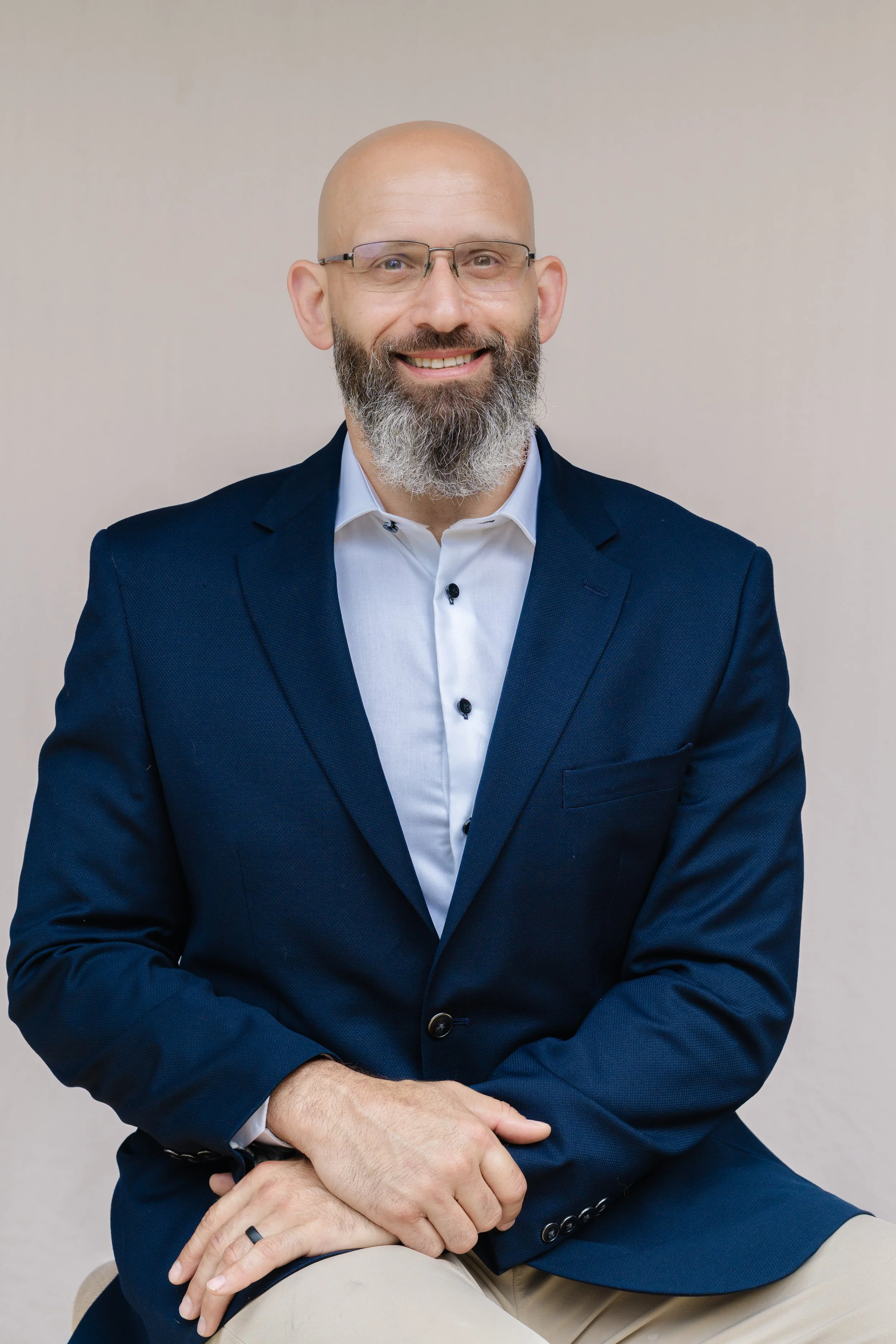 Professional portrait of a man with glasses and a beard in a navy suit, seated