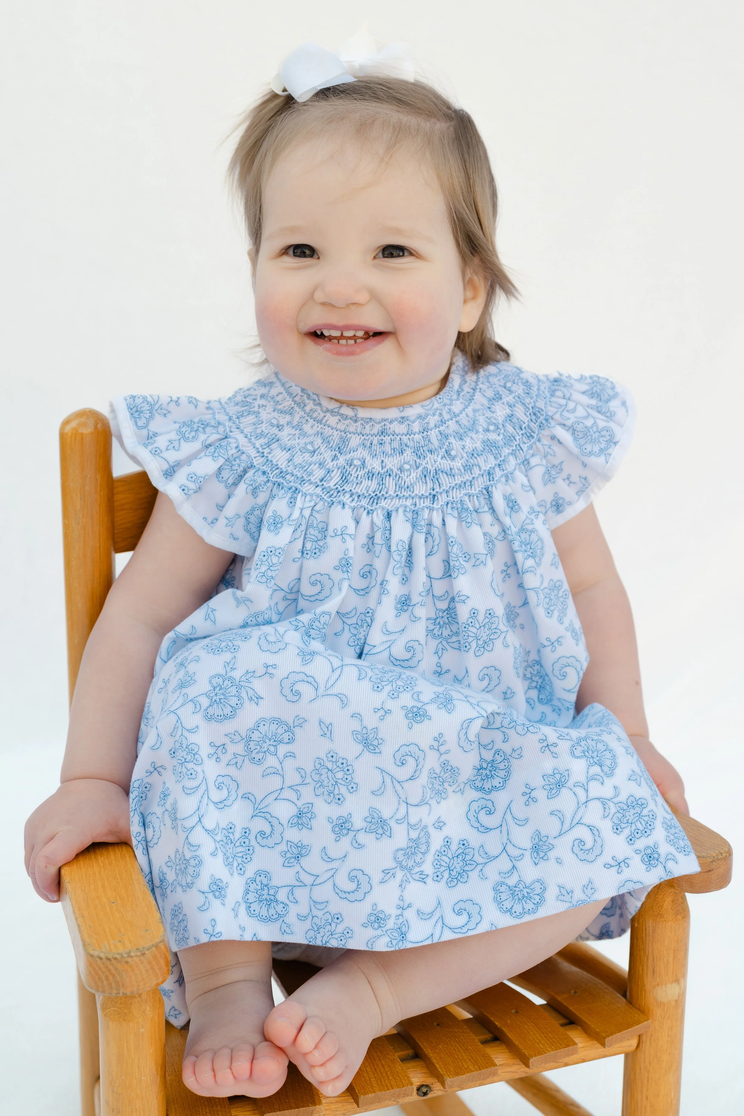 Portrait of a toddler girl in a blue floral smocked dress on a wooden chair