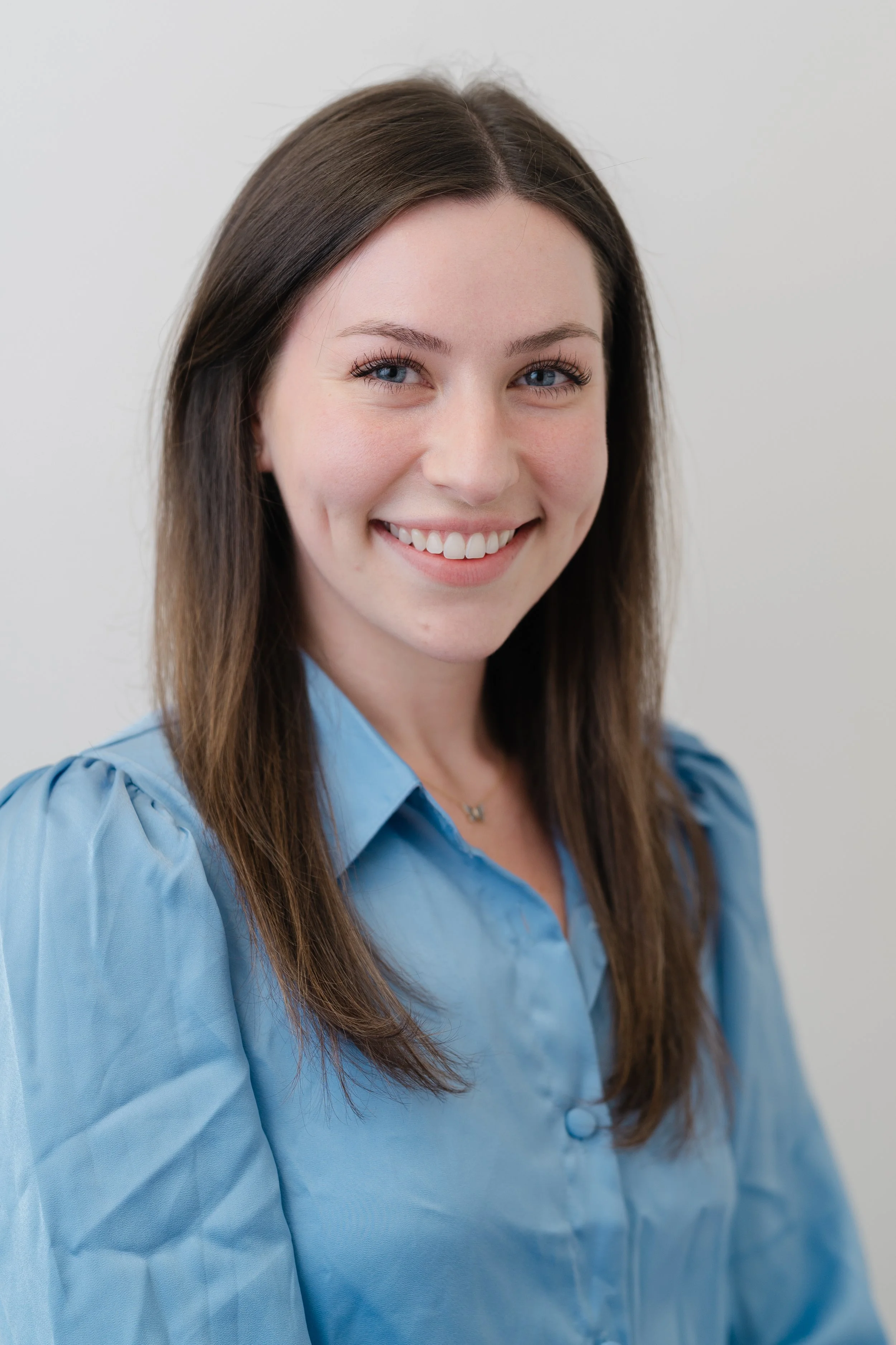 Professional headshot of a smiling woman with long brown hair in a light blue button-up shirt