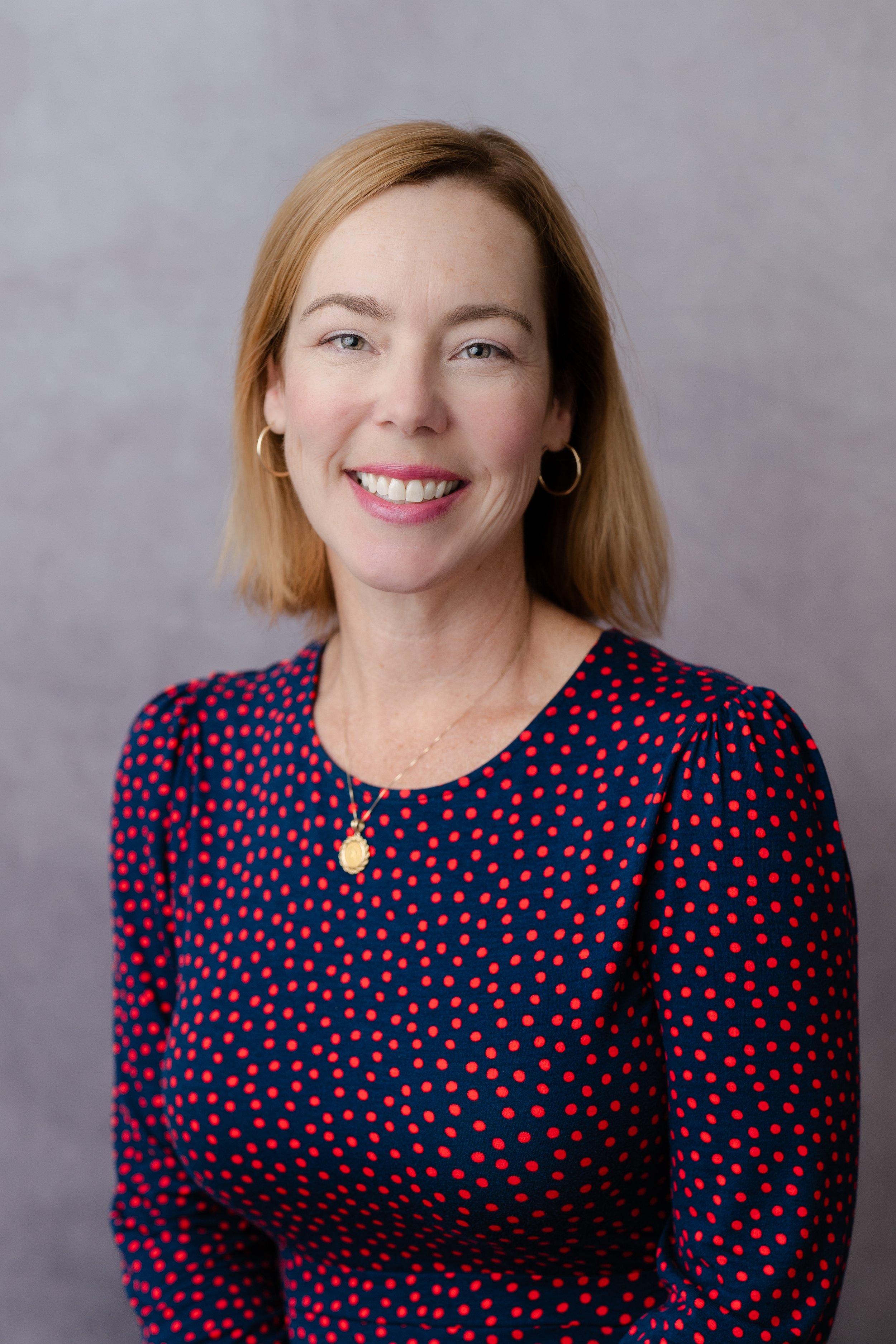 Professional headshot of a smiling woman with auburn hair in a navy polka dot top