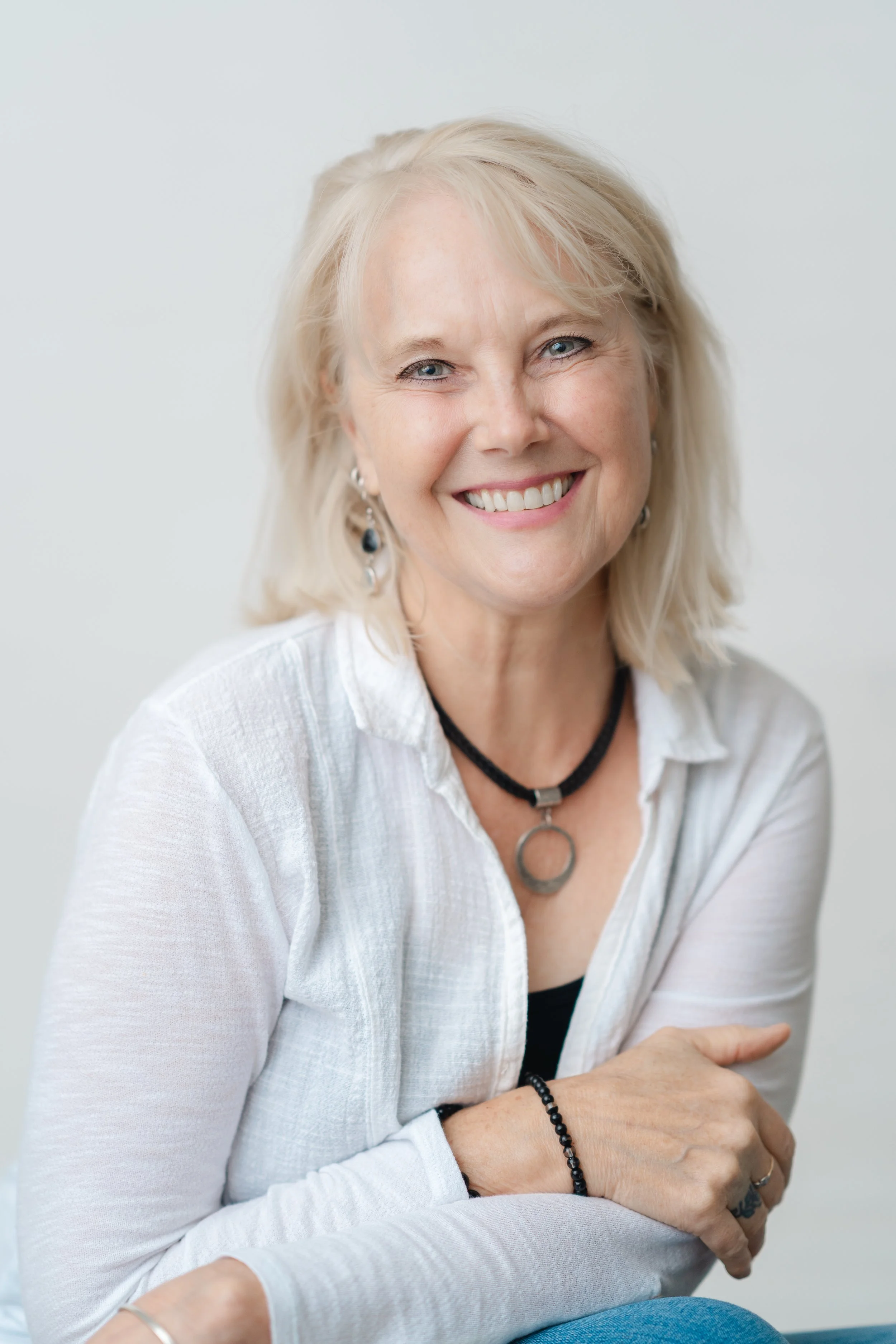Professional headshot of a smiling woman with silver blonde hair and a pendant necklace in a white cardigan