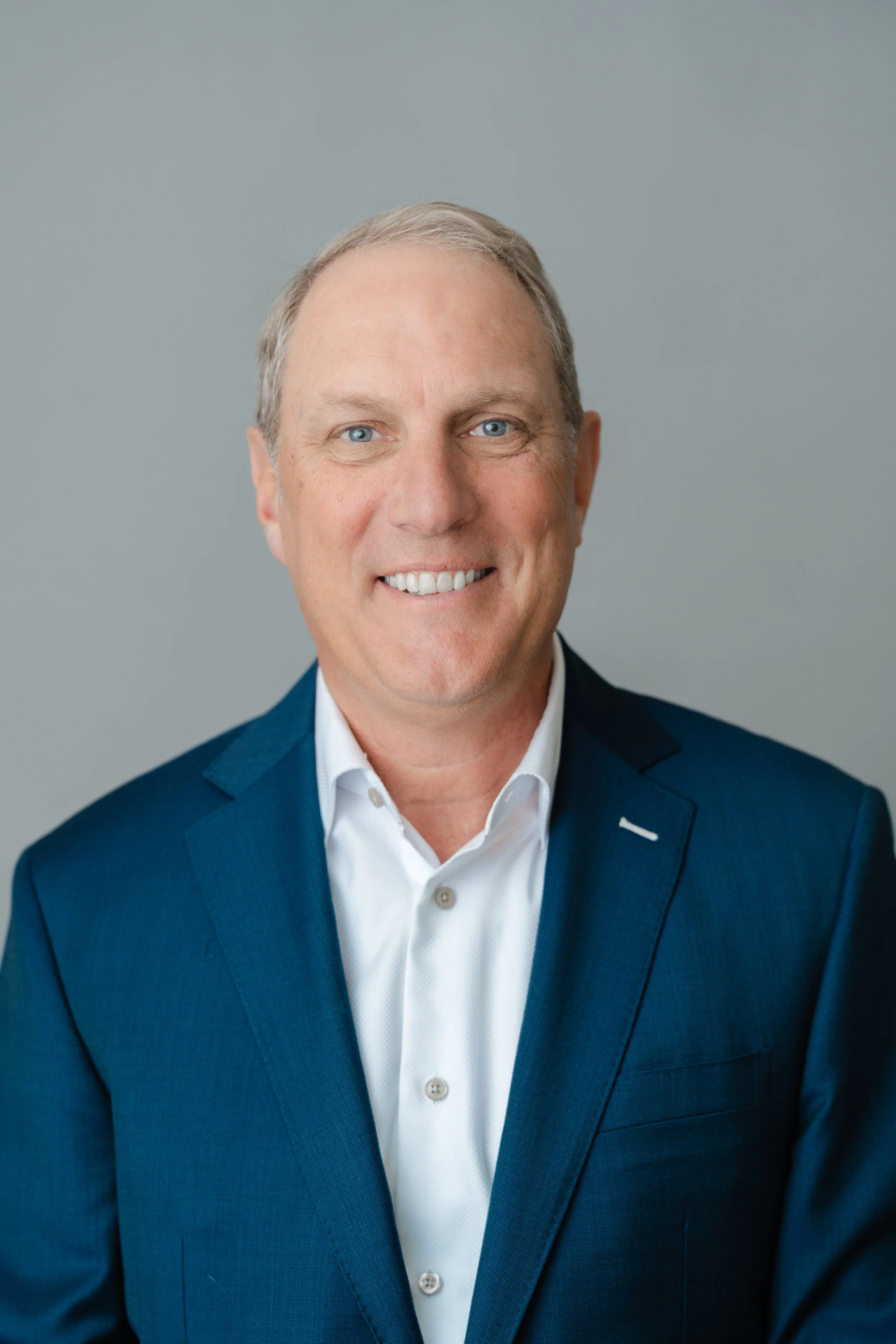Professional headshot of a smiling man with gray hair in a navy blazer and open-collar white shirt