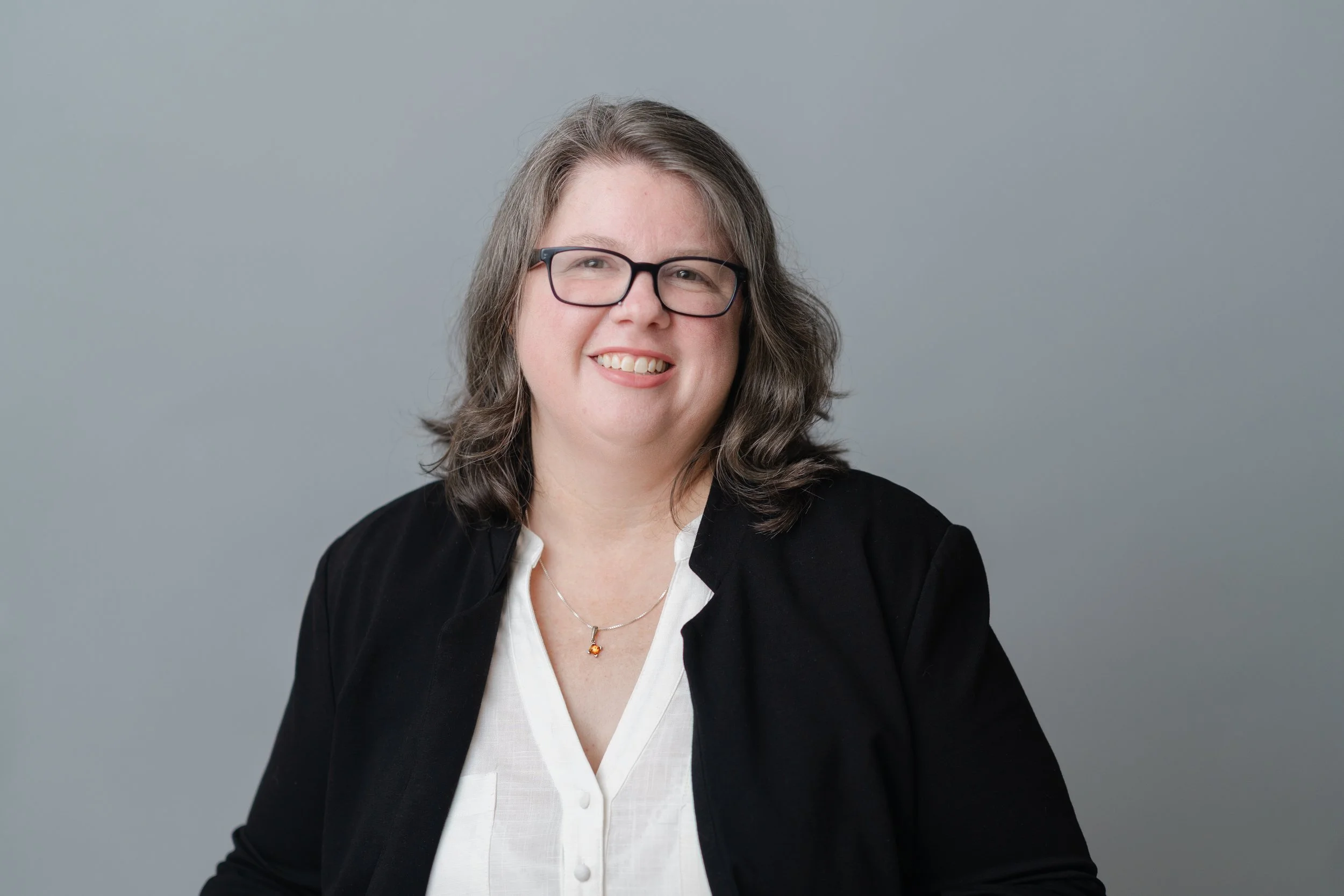 Professional headshot of a woman with glasses and wavy brown hair in a black blazer