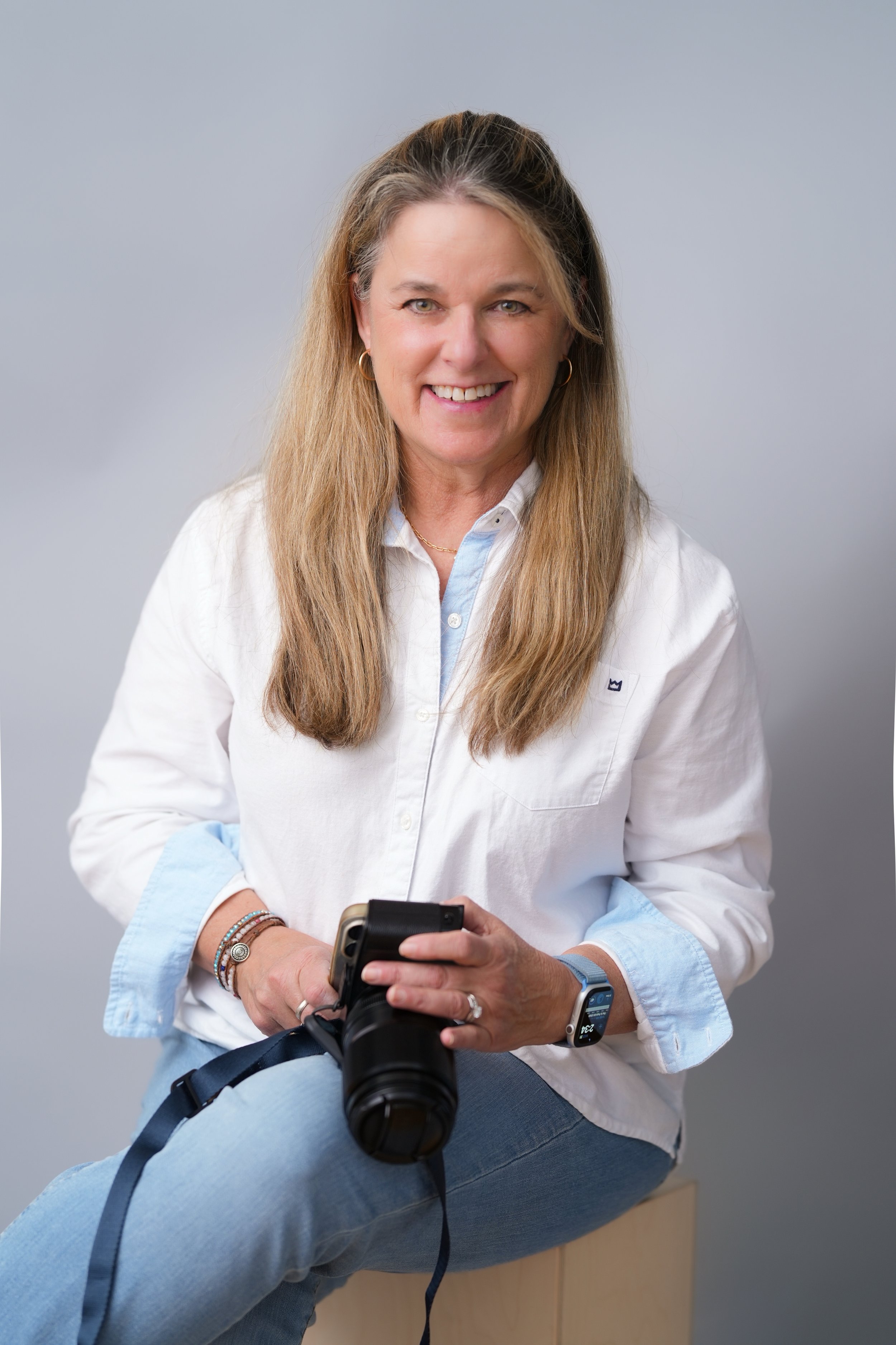 Woman with long light brown hair wearing a white button-down shirt and jeans, sitting with legs crossed, holding a camera, and using a smartphone on a light gray background.
