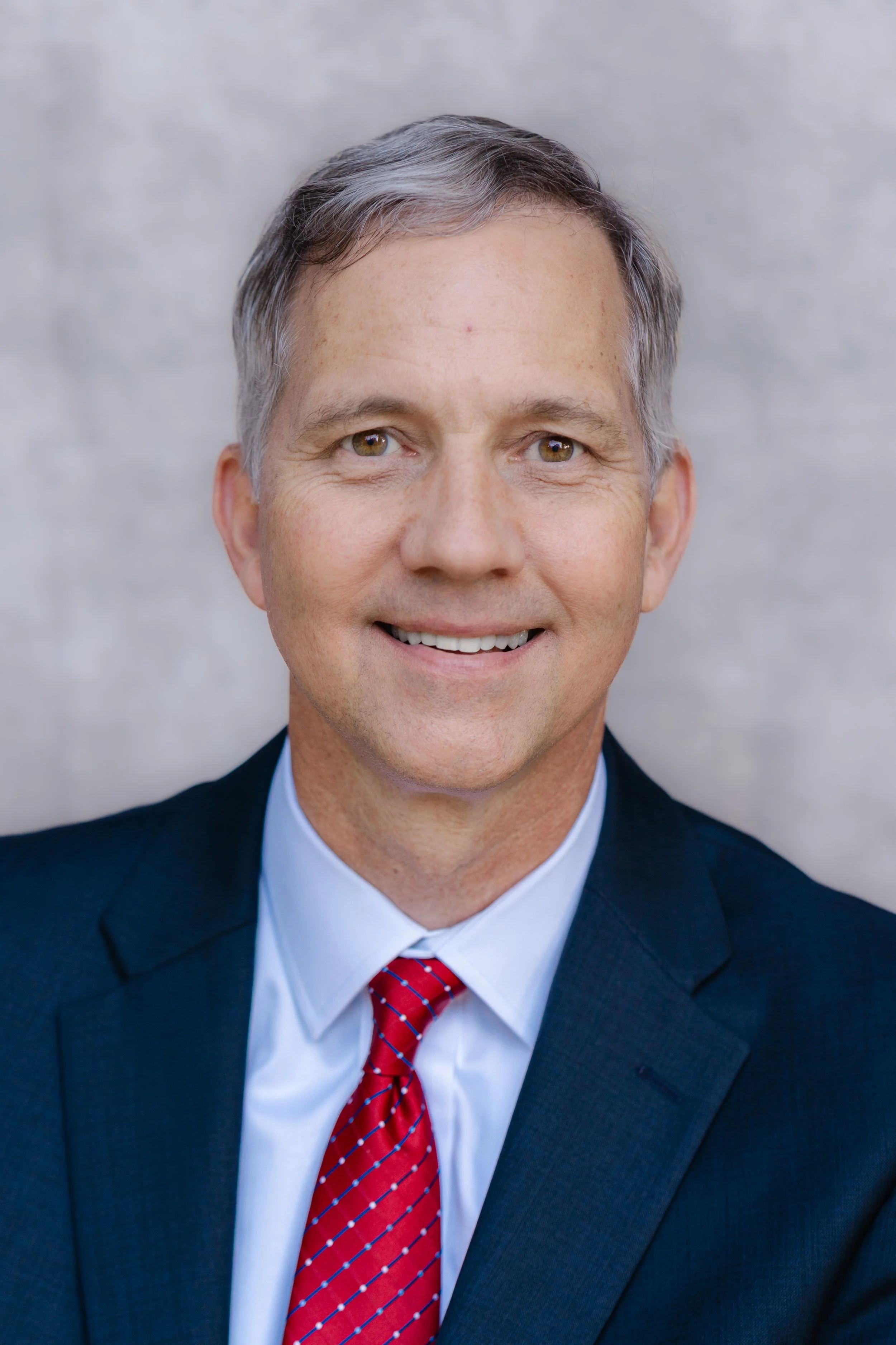 Professional headshot of a smiling man with gray hair in a navy suit and red tie