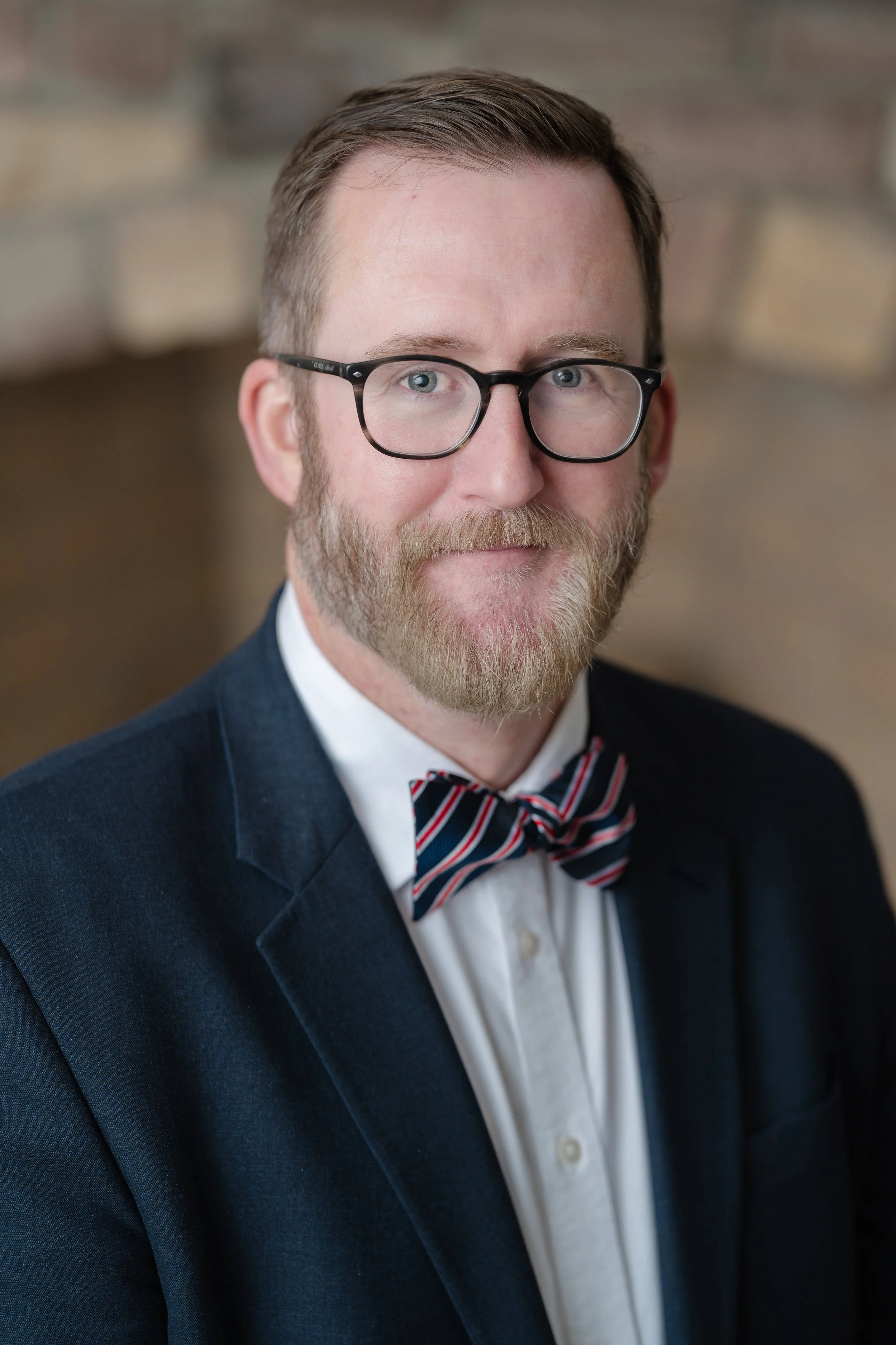 Professional headshot of a man with glasses and a beard in a navy blazer and bow tie
