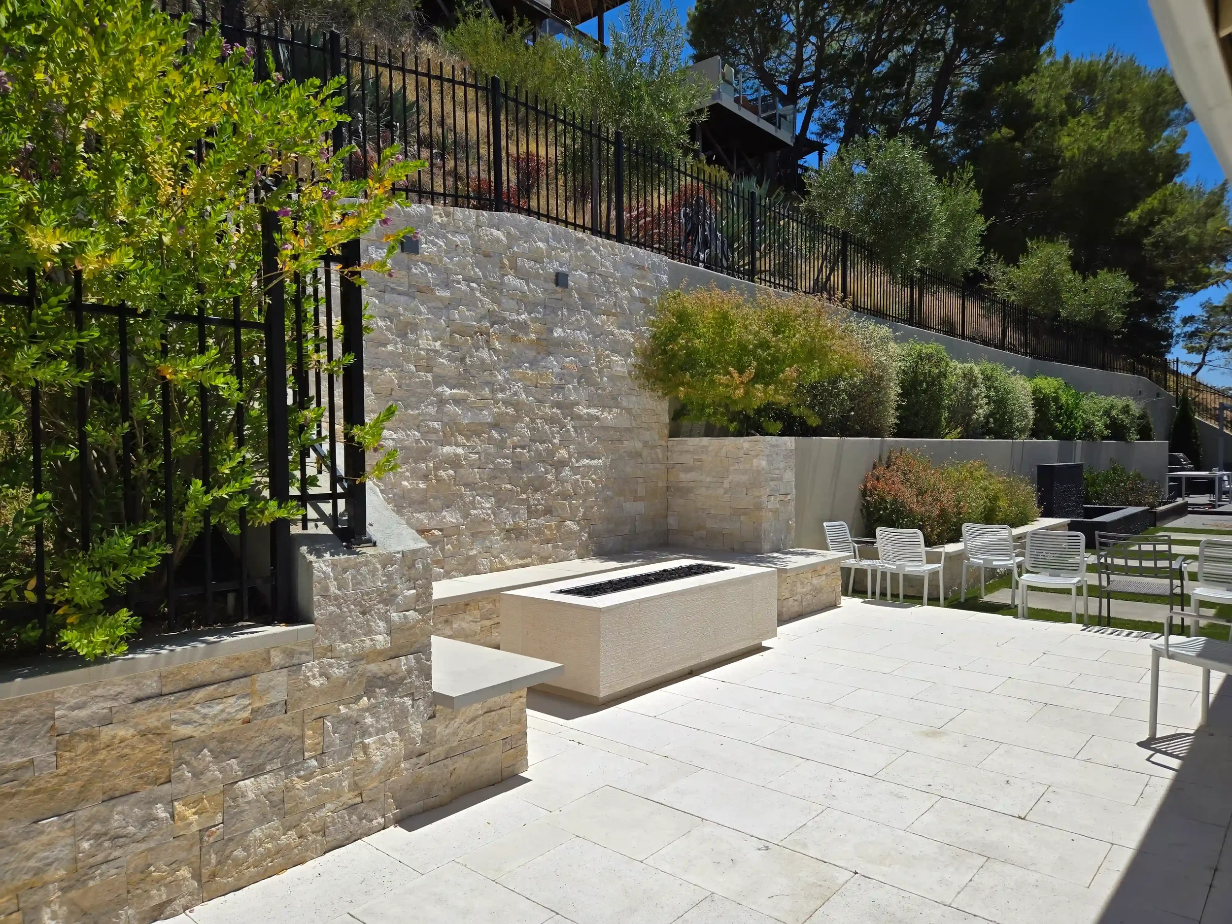 Outdoor modern seating area with white chairs, a stone bench, landscaped garden with bushes, grass, a paved walkway, and a high retaining wall with trees and sky in the background retaining wall and Civil Engineering by Massoudi Consulting engineers.