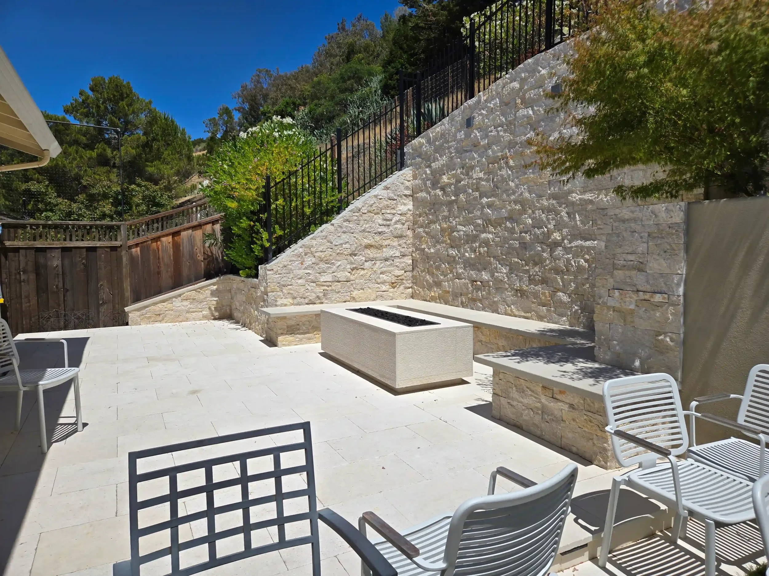 A modern outdoor patio with white tiled flooring, a built-in fire pit, stone wall with a metal railing, and several white chairs; surrounded by trees and a hillside