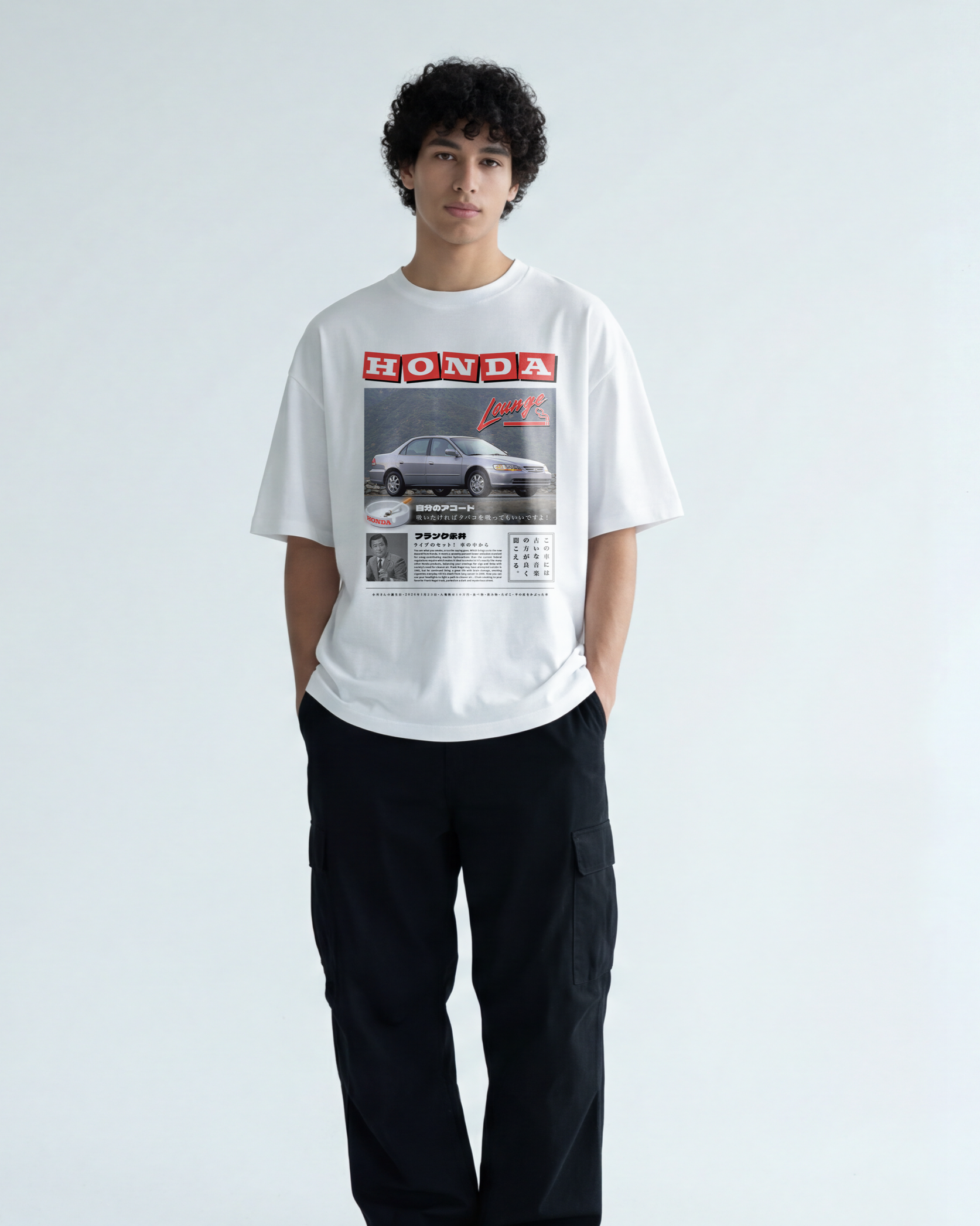 A young man with curly black hair wearing a white T-shirt with a vintage Honda advertisement and black cargo pants, standing in front of a plain light background.