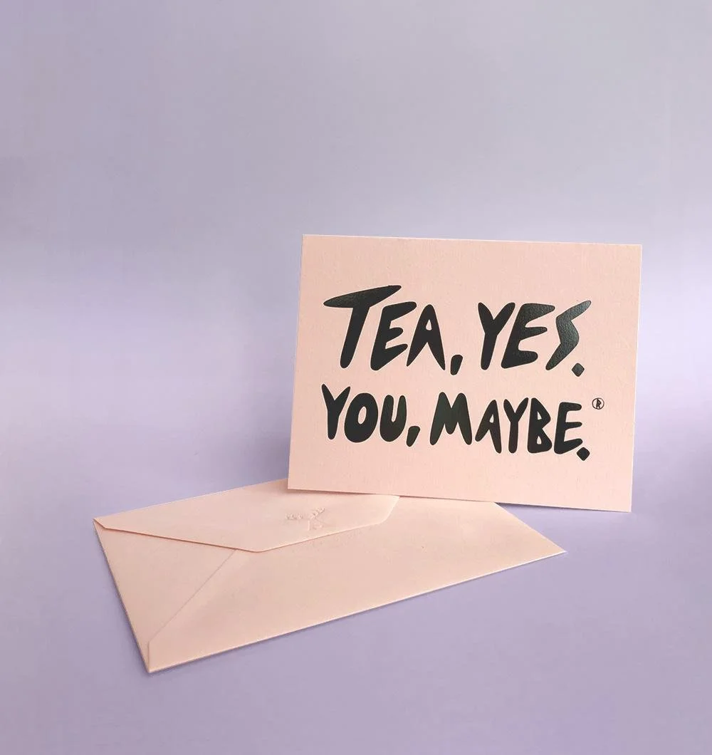 A pink greeting card with playful black text saying 'Tea. Yes. You, maybe.' is standing upright next to a folded pink envelope on a light purple background.