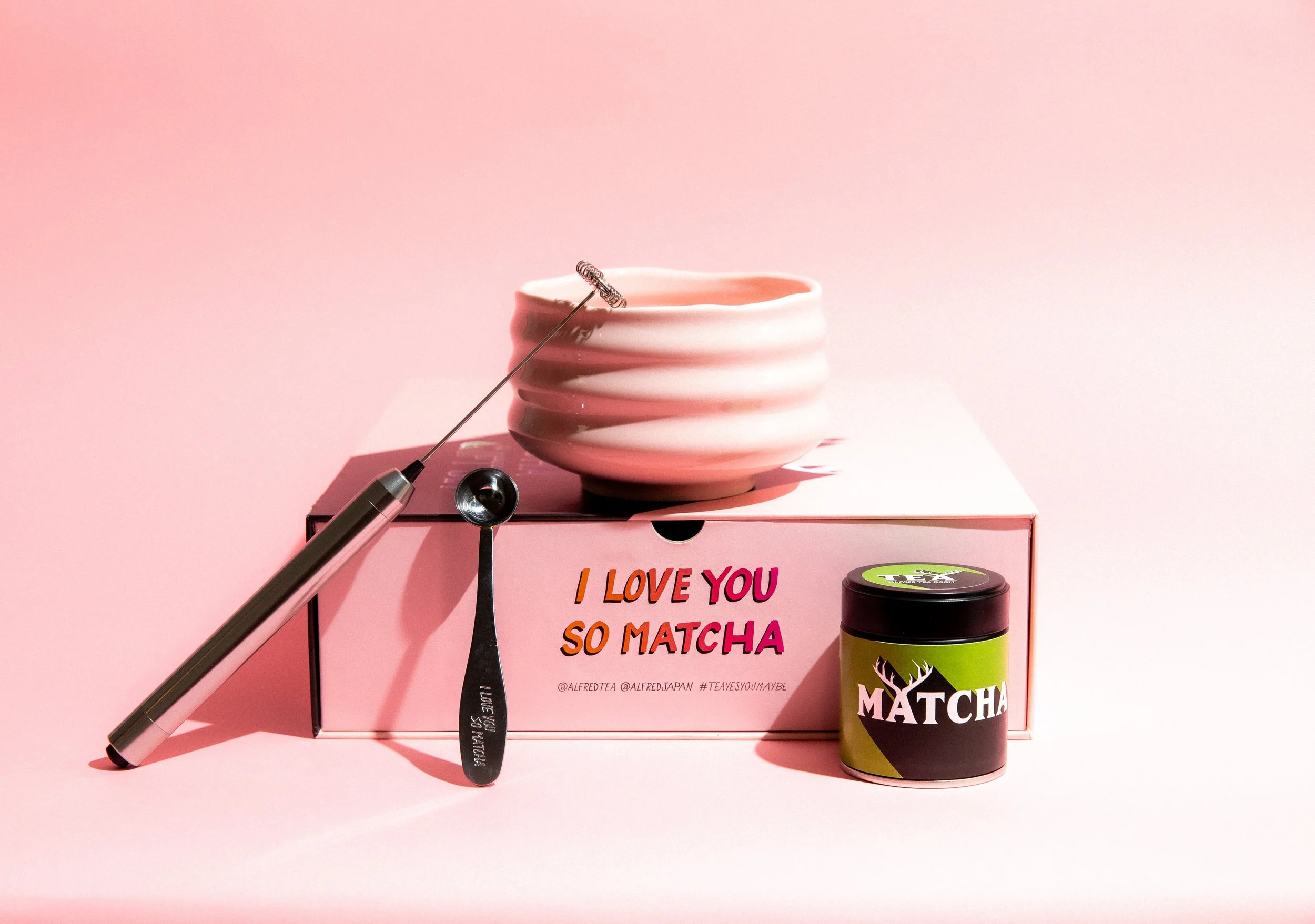 Pink box with printed message, a ceramic bowl, a matcha tea canister, a metal milk frother, and a silicon whisk on top. The background is pink.