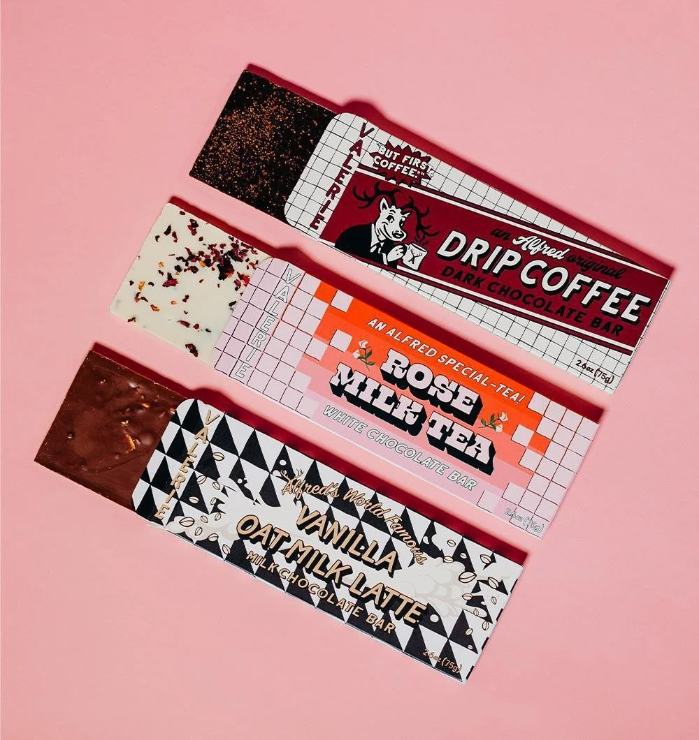 Four chocolate bar wrappers arranged on a pink background, featuring flavors like dark chocolate coffee, white chocolate with rose and millet, vanilla oat milk latte, and a white chocolate bar with colorful sprinkles.