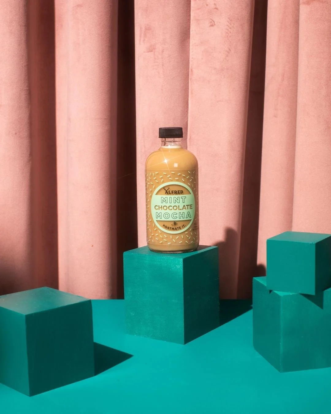 Bottle of Alfred Mint Chocolate Mocha drink placed on teal platforms, with pink curtains in the background.