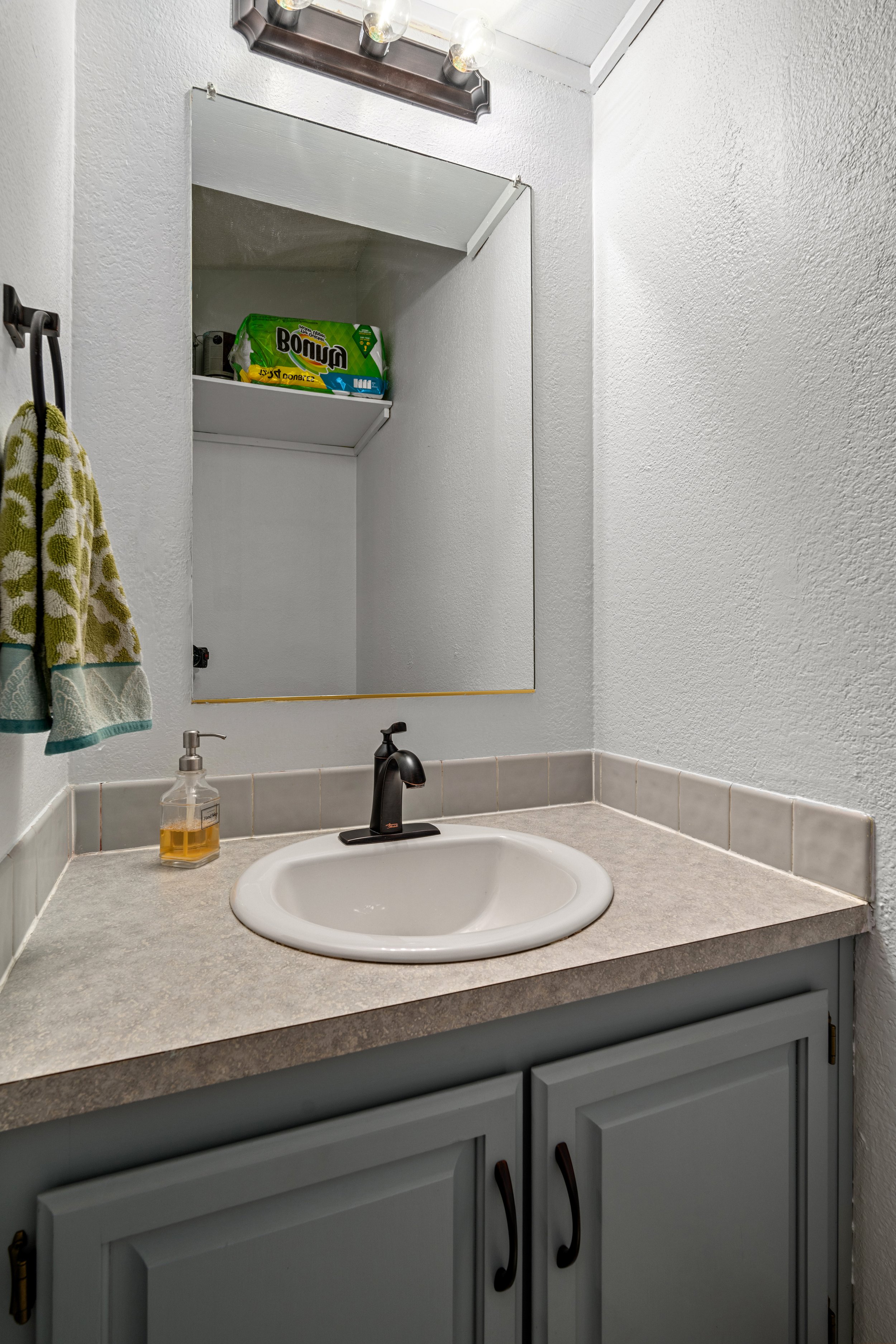 Bathroom sink with mirror and light fixture, green towel on holder, soap dispenser on counter, storage shelf reflected in mirror.