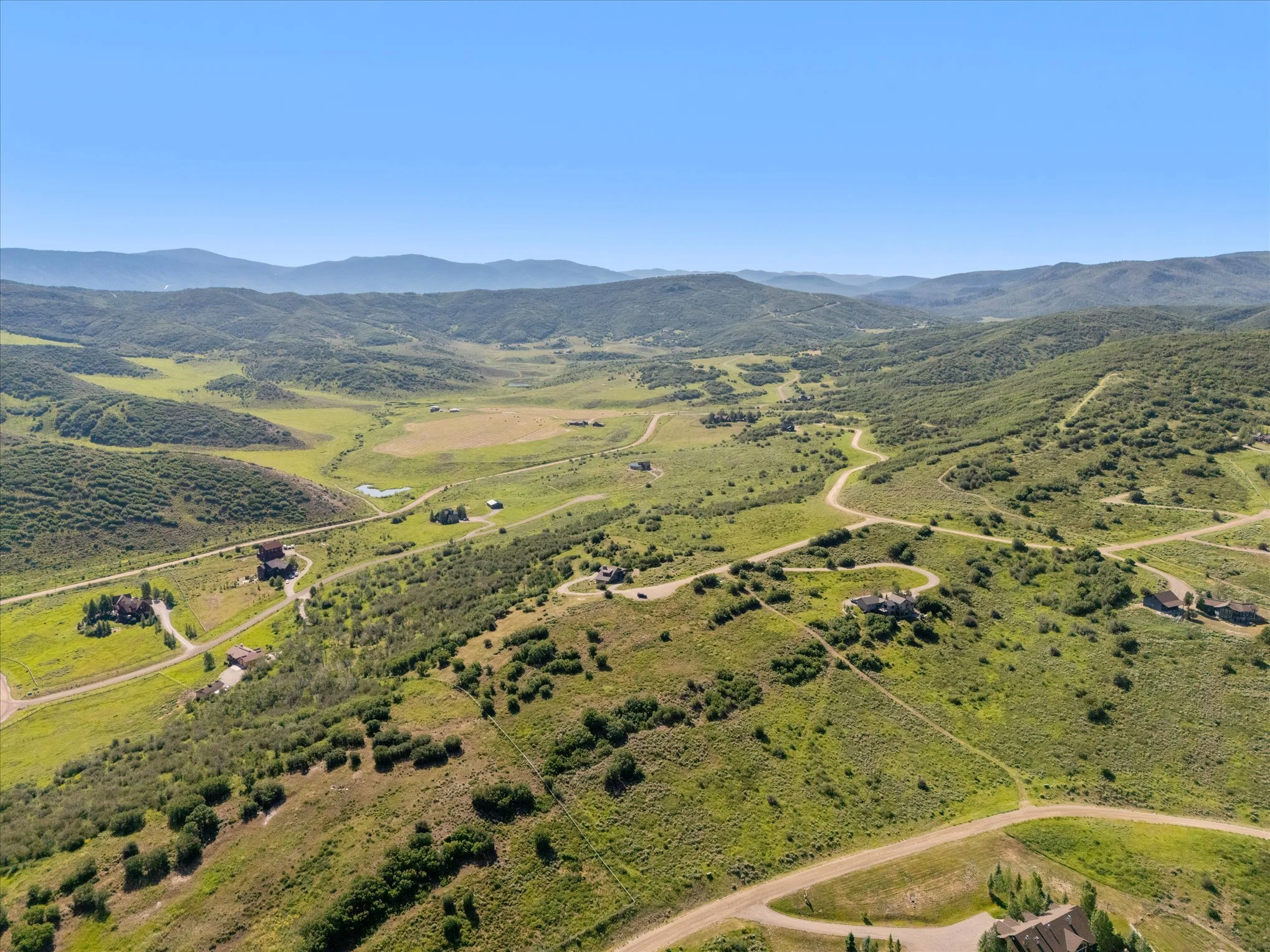 Aerial view of a rural landscape with winding roads, scattered houses, green hills, and distant mountains under a clear blue sky.